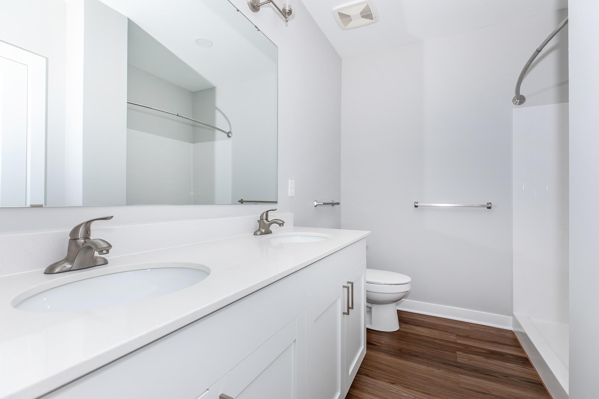 A modern bathroom featuring a double sink with silver faucets, a large mirror, a white toilet, and a walk-in shower with a glass door. The flooring is made of wooden planks, and the walls are a light gray color, creating a clean and spacious appearance.