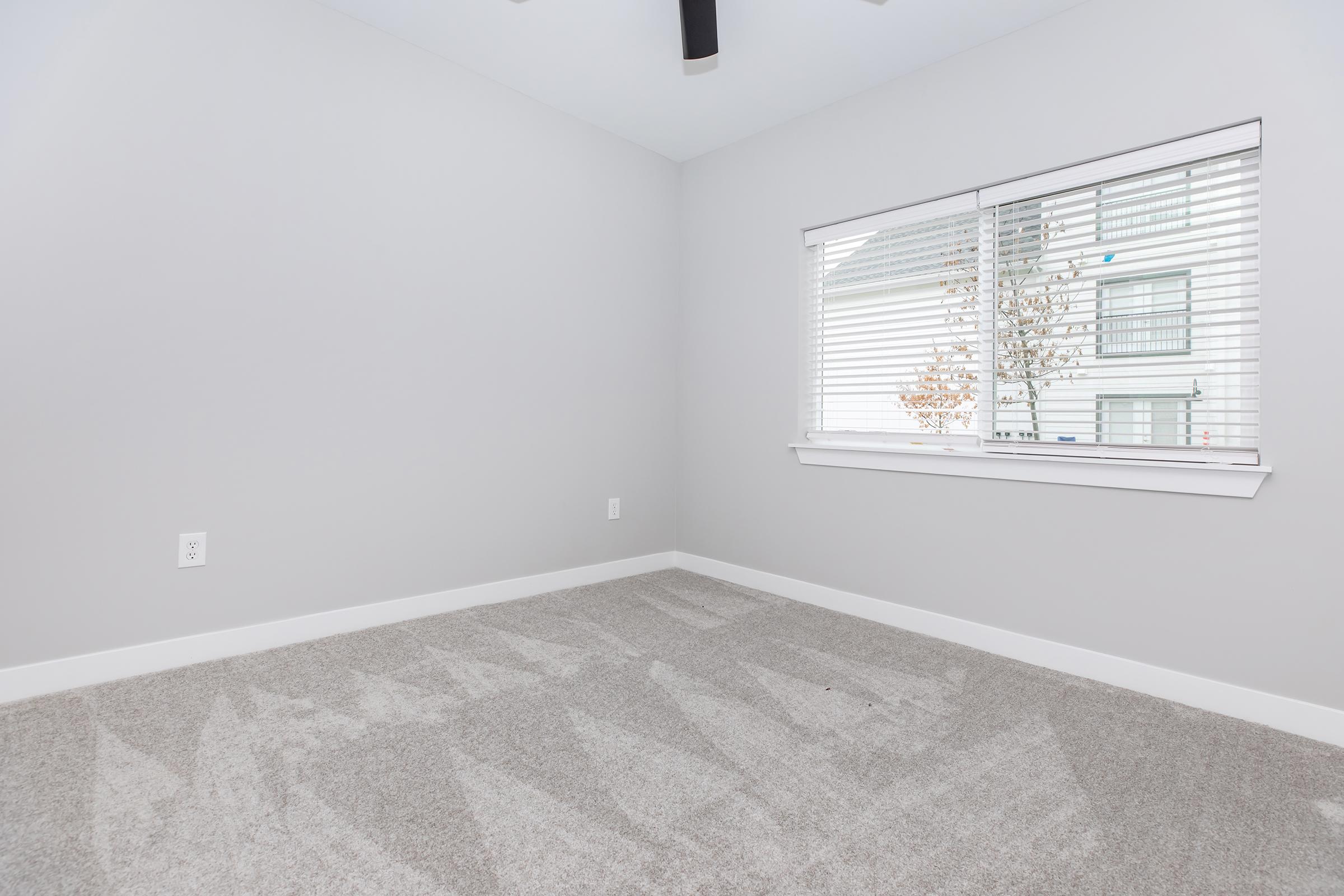 A vacant room featuring gray walls, a ceiling fan, and a large window with blinds. The floor is covered in light-colored carpet, and the space is well-lit with natural light entering through the window. The atmosphere is minimalistic and uncluttered.