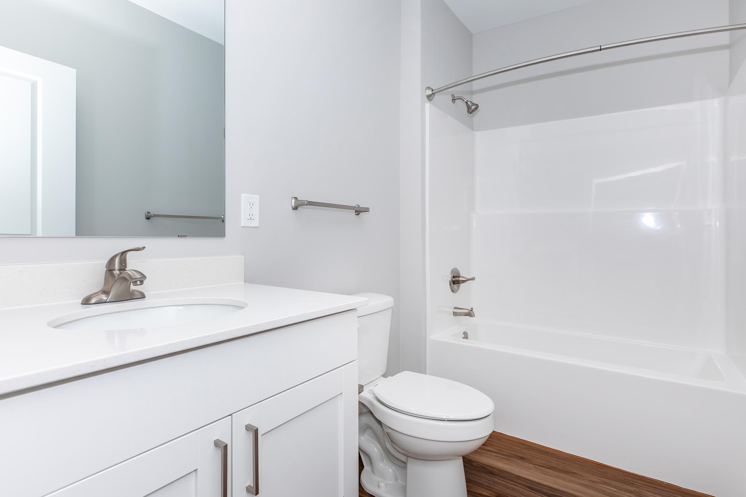 A modern bathroom featuring a clean white bathtub, a toilet, and a sink with a sleek countertop. The walls are painted light gray, and there's a large mirror above the sink. The flooring is a warm wood tone, enhancing the contemporary design.