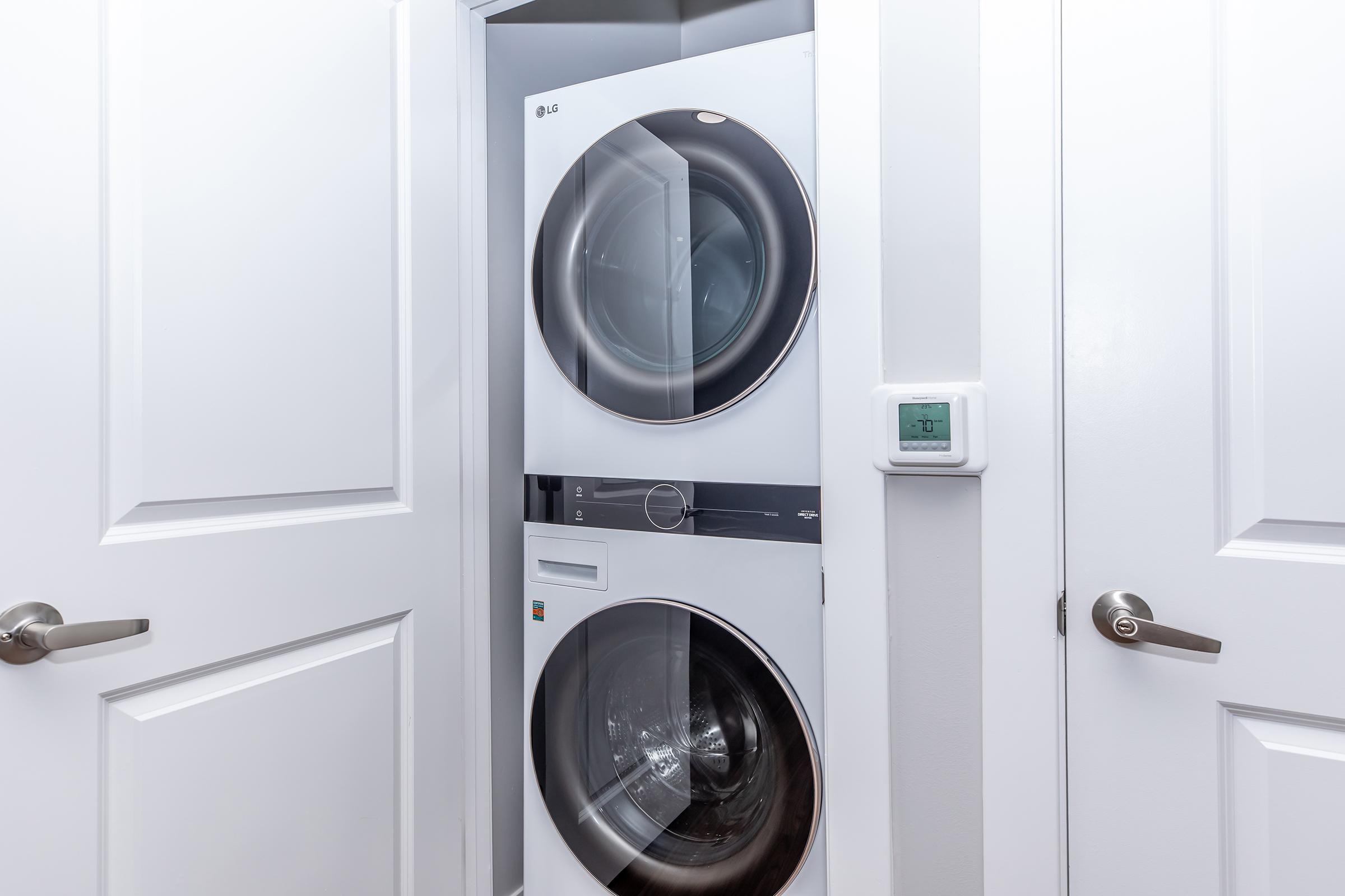 A stacked LG washer and dryer unit in a laundry closet, with white doors and a digital thermostat on the wall beside it. The doors of the closet are closed, and the appliances are modern and sleek in design.
