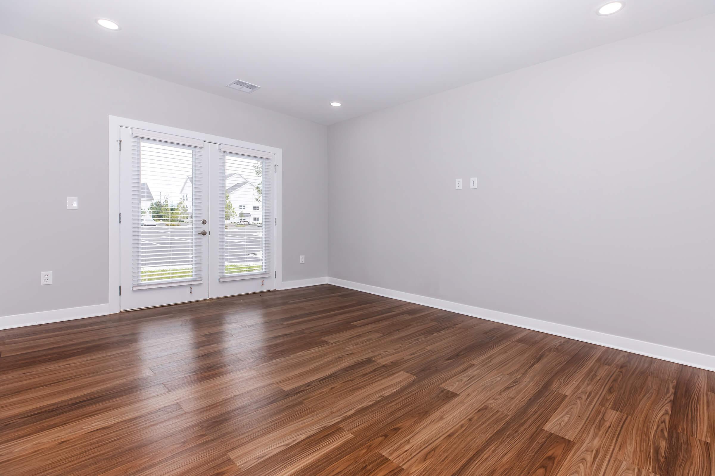 A spacious, empty room with light gray walls and wooden flooring. Two large windows with blinds provide natural light. The room features a set of double doors leading outside and has no furniture or decorations, creating a minimalist look.