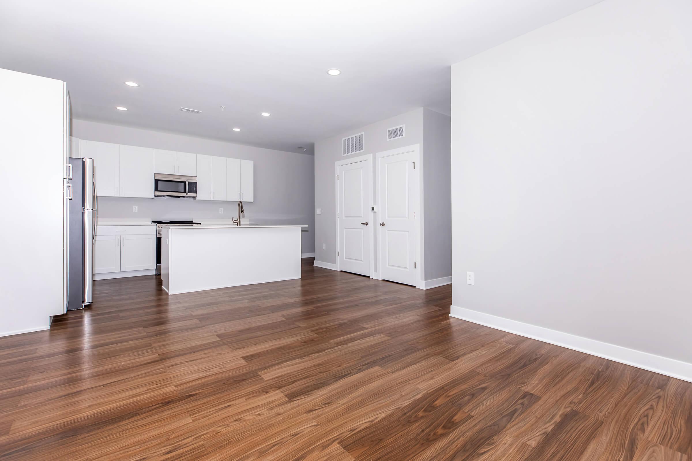 Spacious and modern open-concept kitchen and living area. The kitchen features white cabinets, stainless steel appliances, and a large island with a clean countertop. The flooring is dark wood, and the walls are painted light gray, creating a bright and airy atmosphere. Two closed doors are visible, leading to additional rooms.