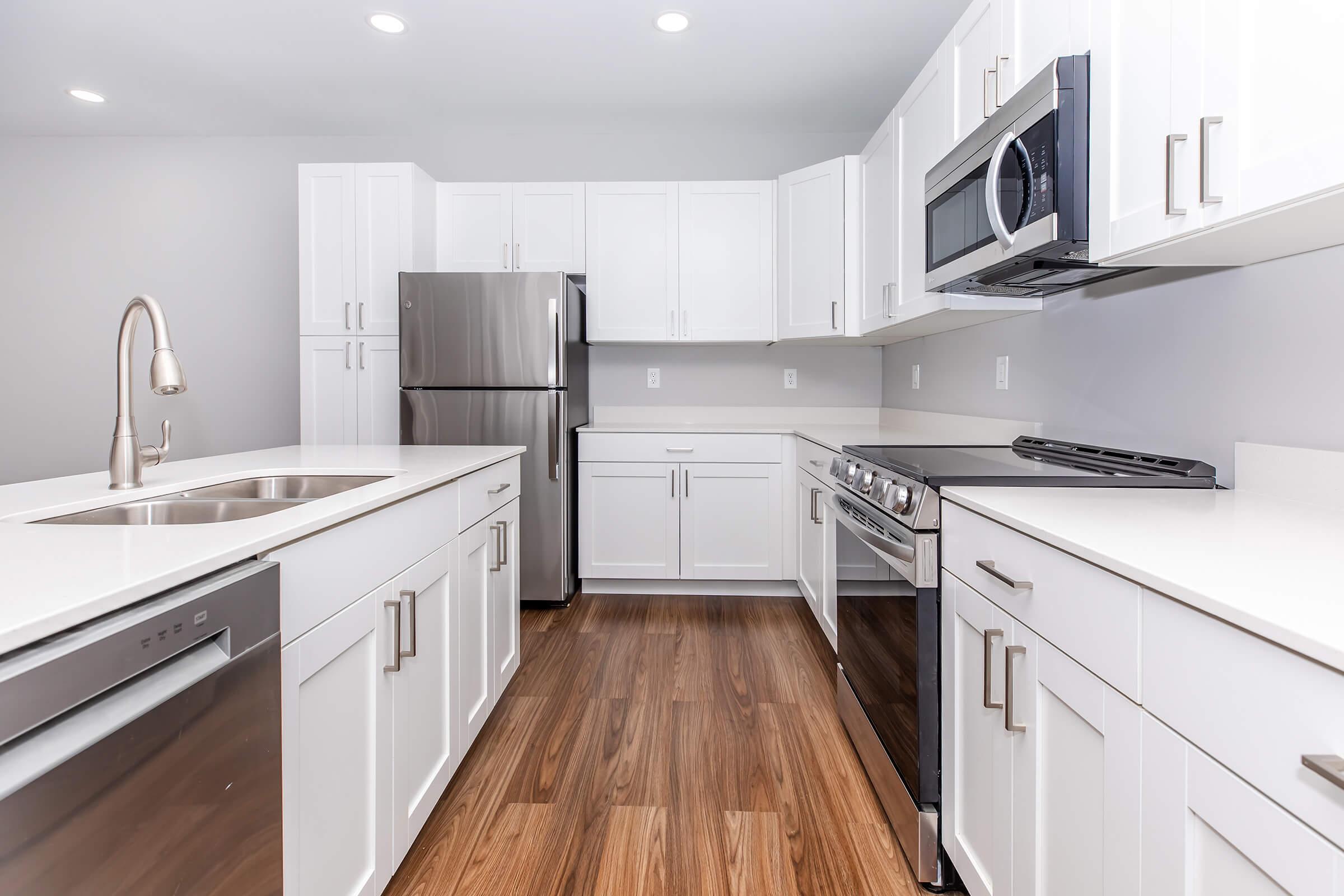 Modern kitchen featuring white cabinetry, stainless steel appliances, a double sink, and a sleek countertop. The space has a light color scheme with wooden flooring and ample natural light from ceiling fixtures.