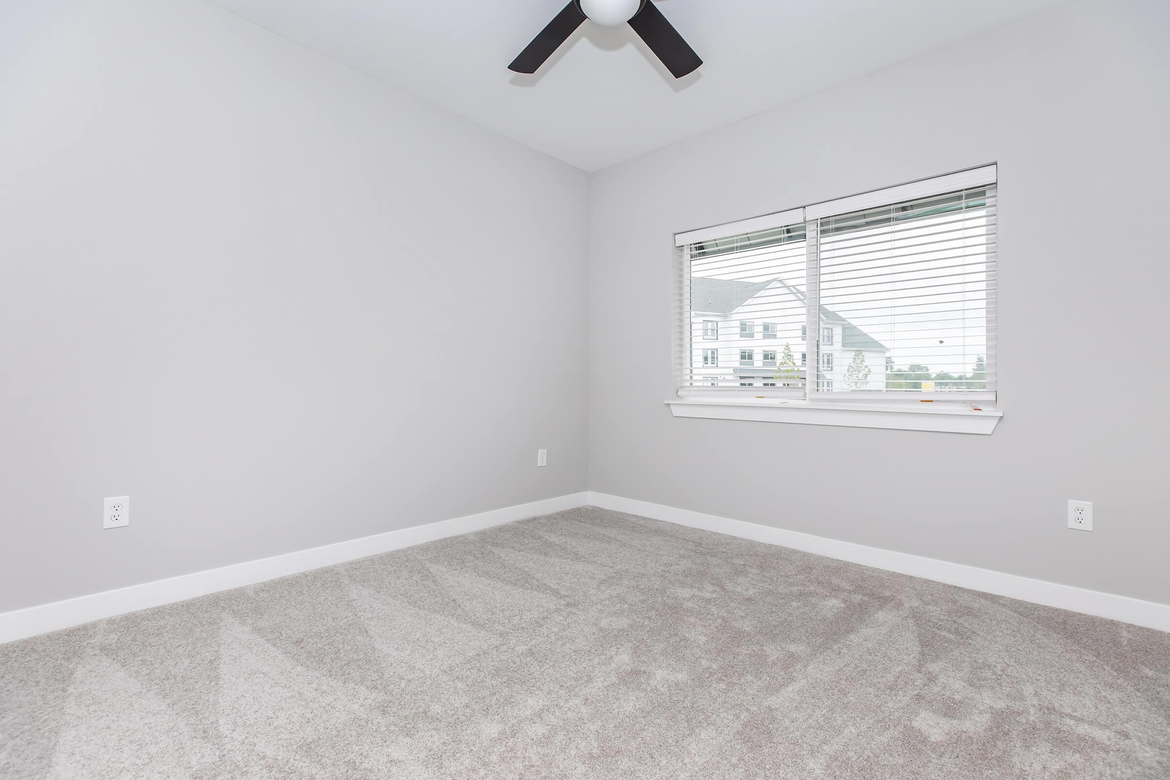 A minimally furnished room with light gray walls and a ceiling fan. The floor is covered in soft, beige carpet. A large window with white blinds allows natural light to enter, revealing a glimpse of an exterior building. The room is clean and spacious, offering a blank canvas for decoration.