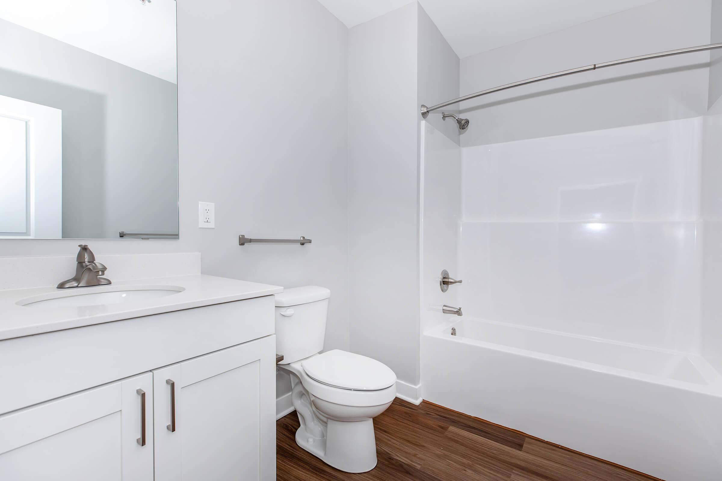 A modern bathroom featuring a white toilet, a bathtub with a glass shower rod, and a single sink vanity with a mirror. The walls are painted light gray, and the flooring is made of wood-like tiles, creating a clean and contemporary look.