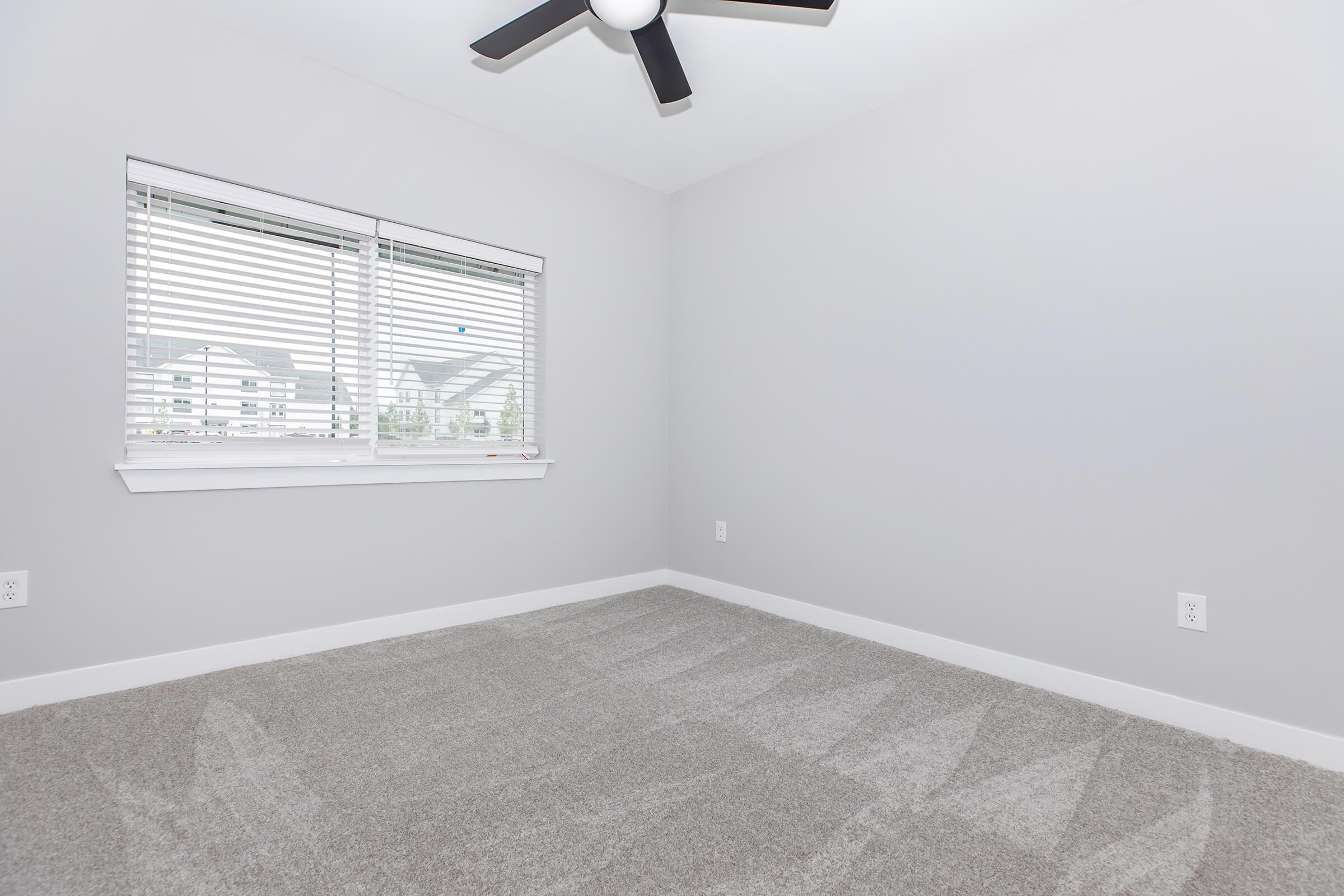 A clean, empty room featuring light gray walls, a large window with white blinds, a ceiling fan, and soft, light gray carpet flooring. The room has a modern feel with plenty of natural light and no furniture. The overall atmosphere is bright and inviting.