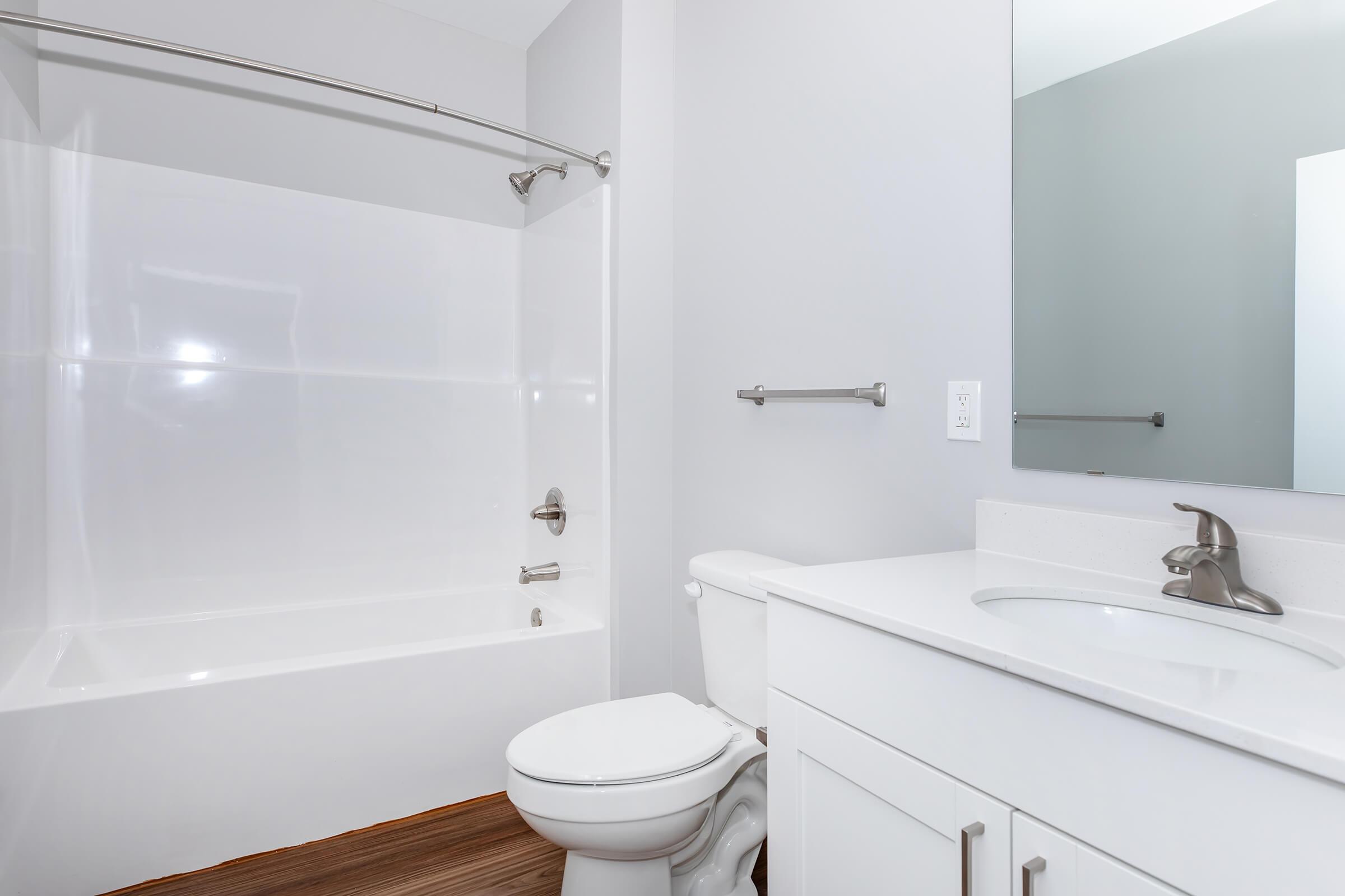 A modern bathroom featuring a white shower tub combination, a white toilet, and a white vanity with a sink. The walls are painted light gray, and there is a large mirror above the sink. The floor is made of brown wood-like material, creating a clean and contemporary look.