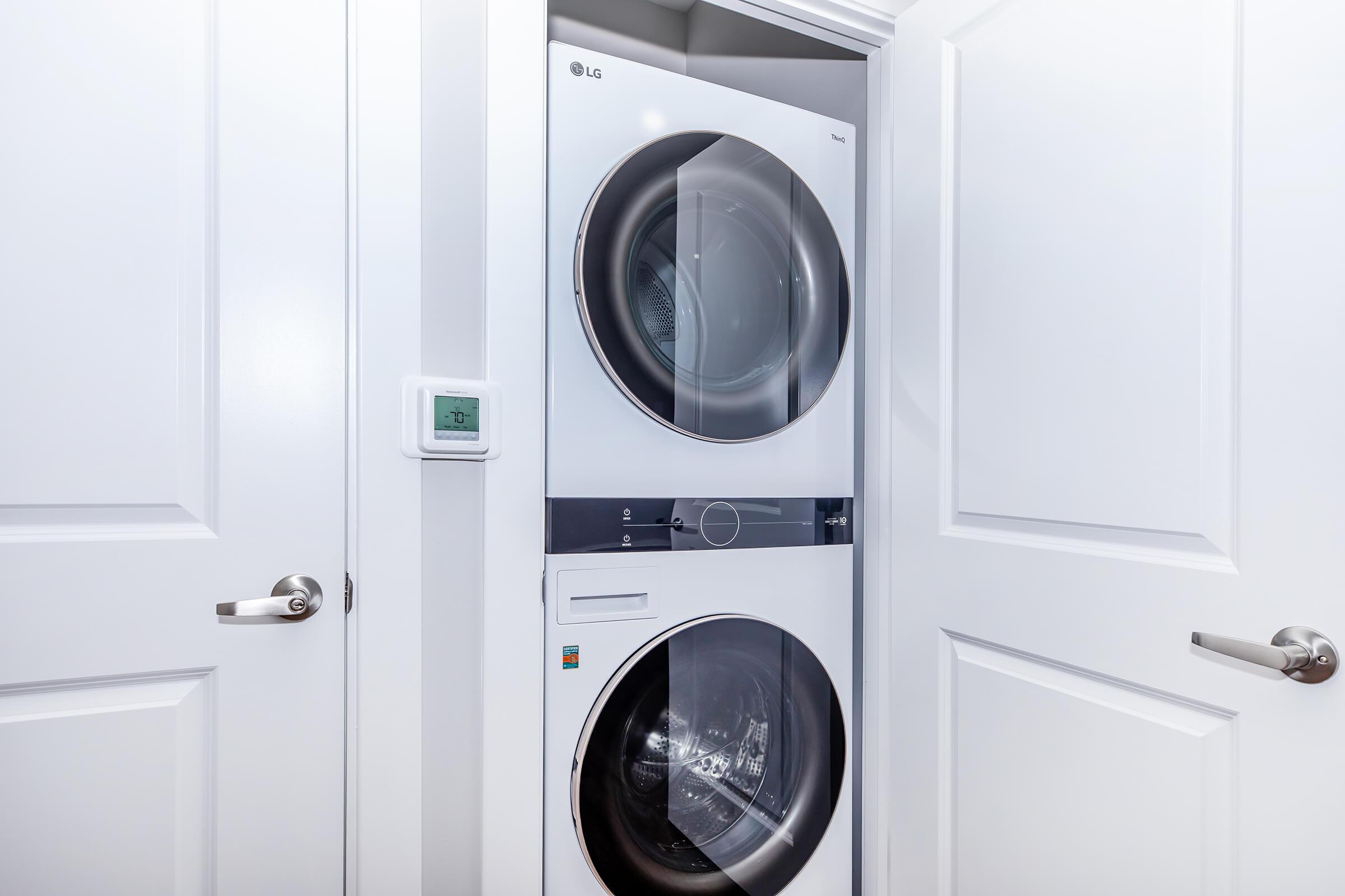 A stacked washer and dryer set in a small laundry closet, with white doors partially open. A digital thermostat is visible on the wall beside the appliances. The design is modern and clean, featuring smooth surfaces and a sleek look.