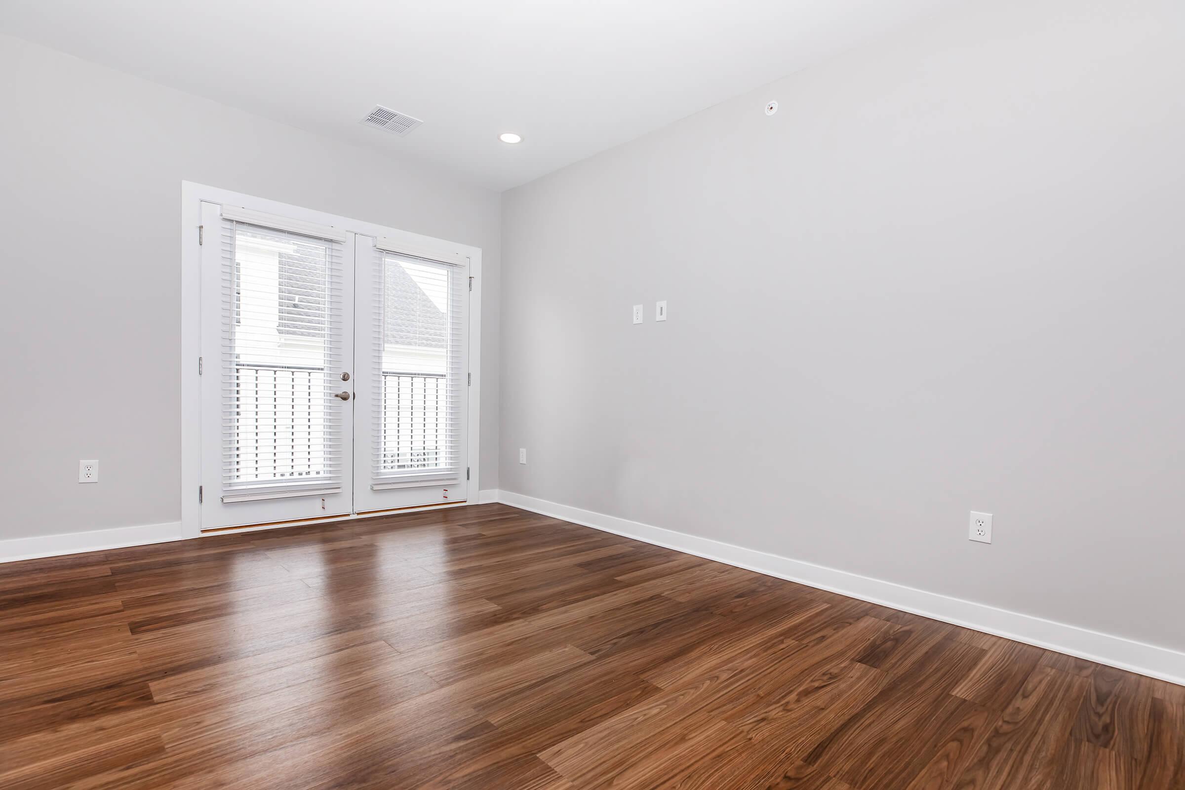 Empty room with light gray walls, hardwood flooring, and large double doors with white shutters leading to a balcony. Two electrical outlets are visible on the wall. The room is well-lit by natural light coming through the doors.