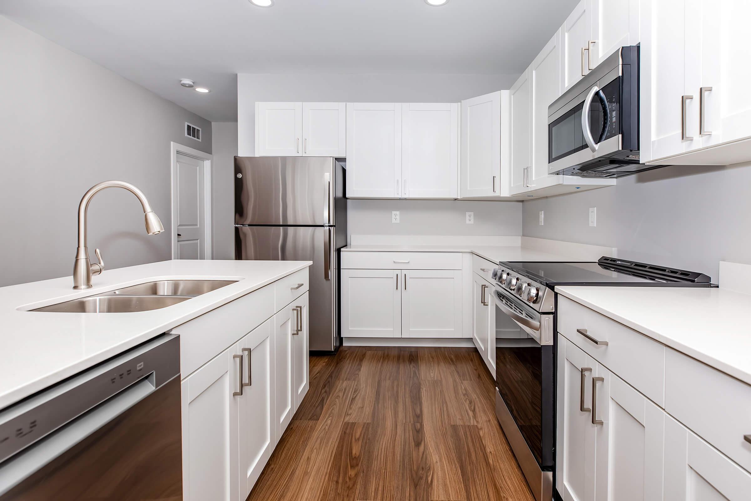 Modern kitchen featuring white cabinetry, stainless steel appliances including a refrigerator, microwave, and oven. The countertop is sleek and light-colored, with a stainless steel sink and a stylish faucet. The floor showcases warm wood tones, creating a clean and inviting atmosphere.