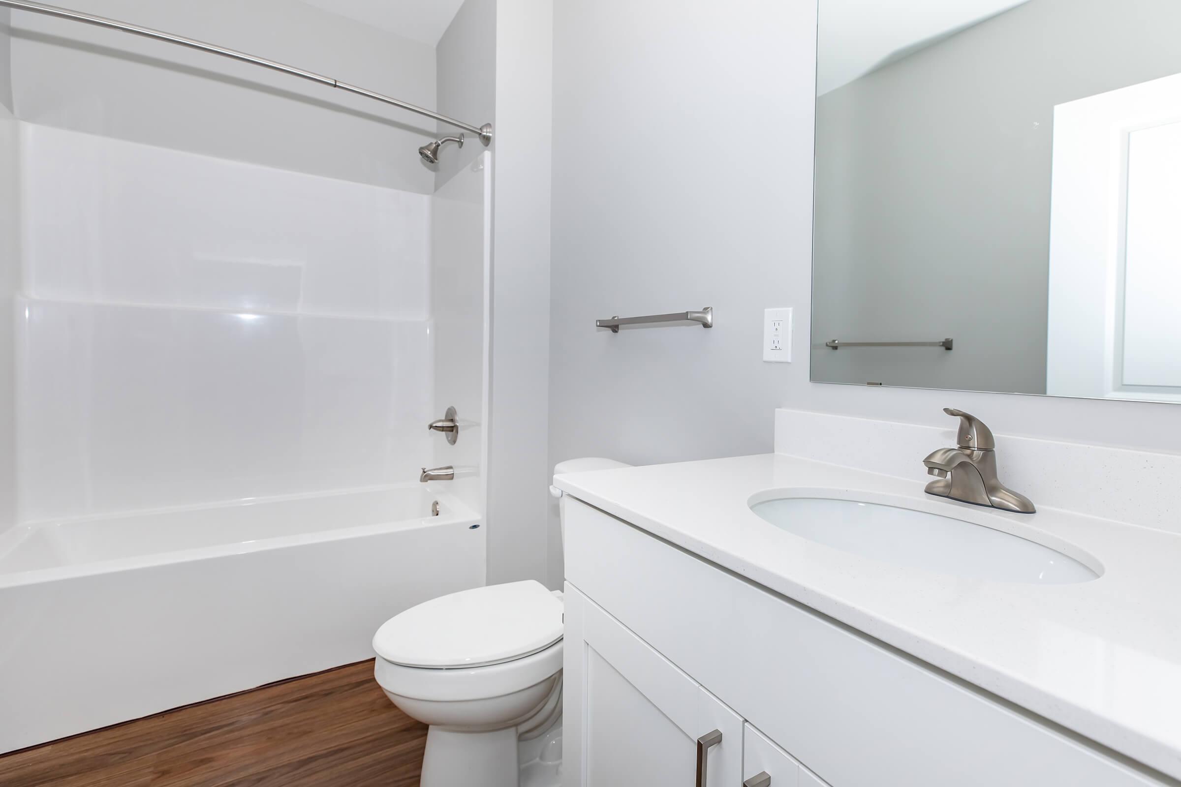 Modern bathroom featuring a white bathtub, a sleek vanity with a sink, a toilet, and a large mirror. The walls are painted in a light gray color, and the floor consists of warm wooden tones. The overall design is clean and minimalistic.