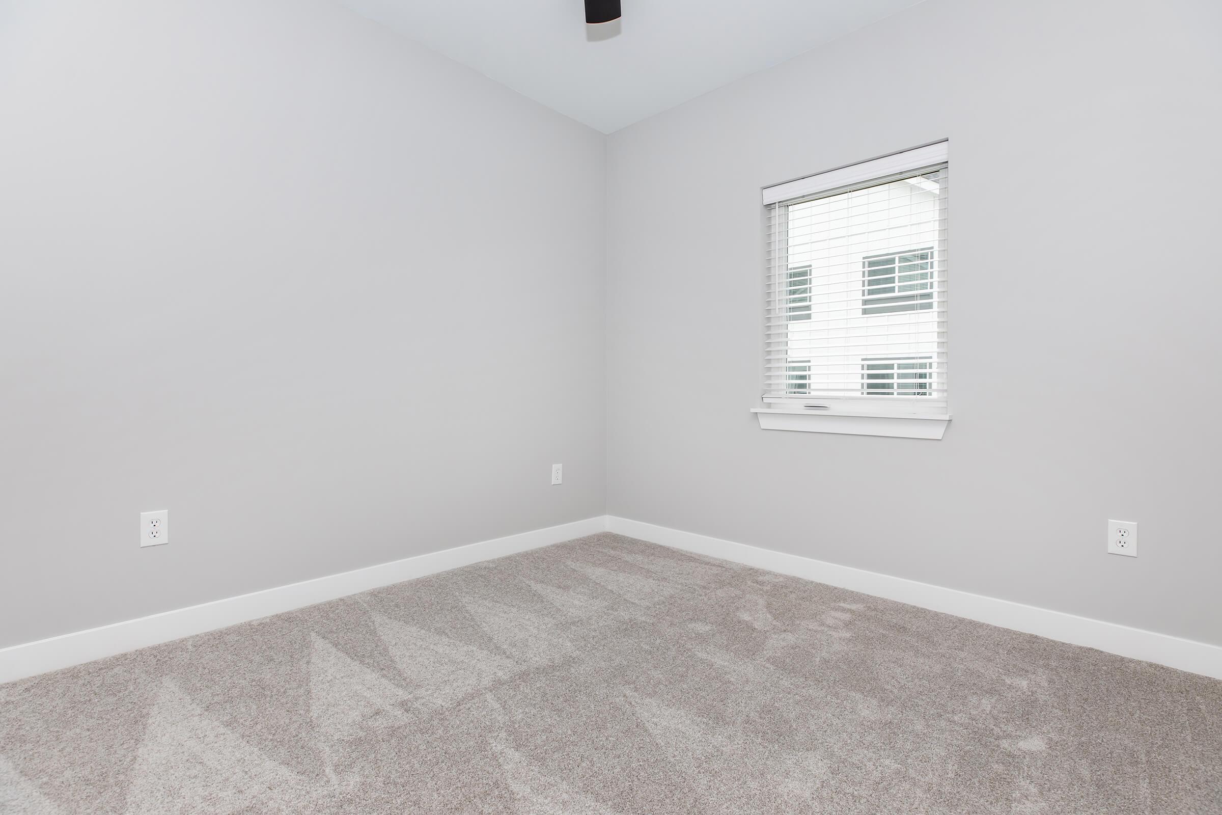 Empty room with light gray walls and carpet. A window with white blinds allows natural light to enter, casting soft shadows on the floor. The space appears modern and minimalistic, featuring a ceiling fan.