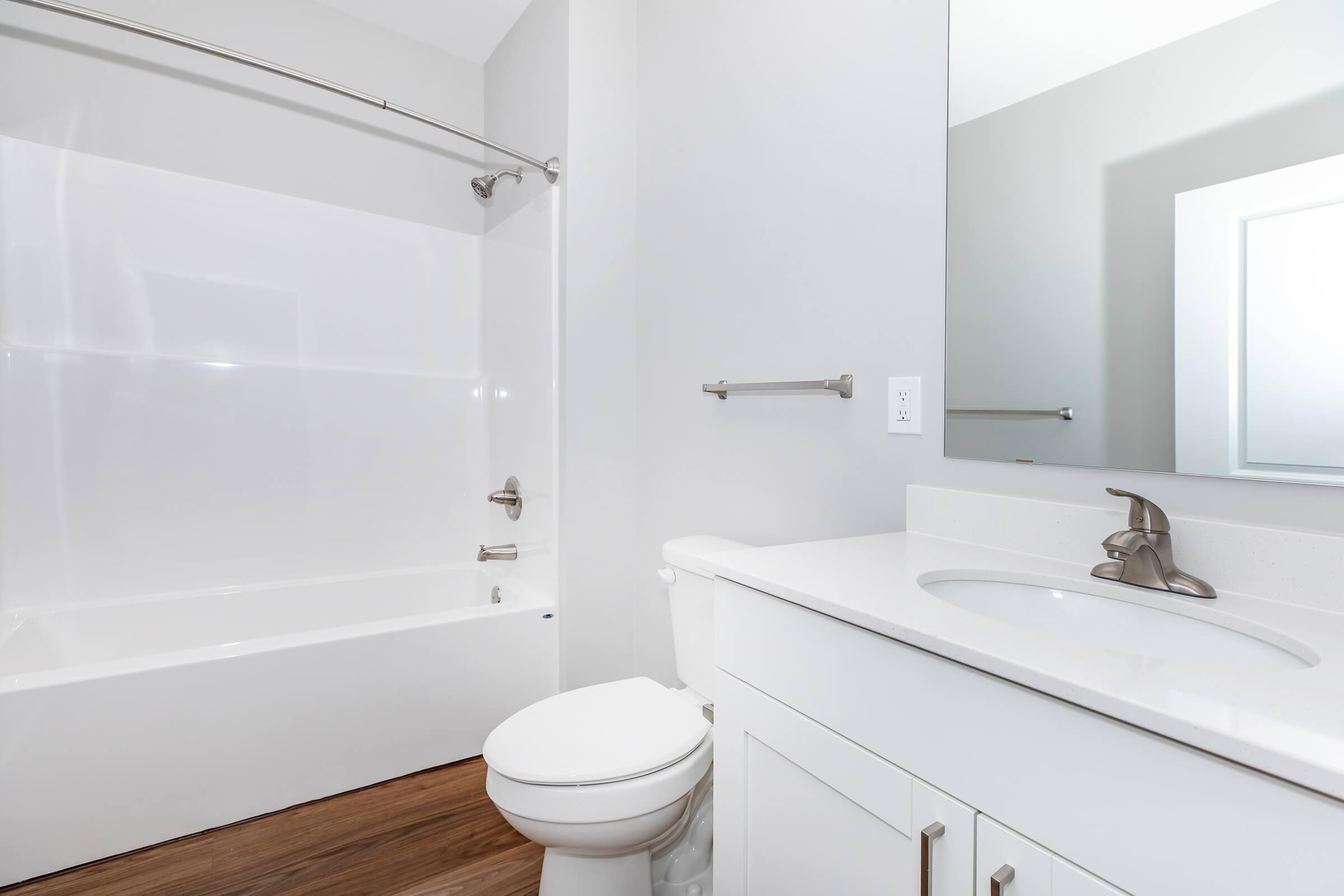 A clean and modern bathroom featuring a white bathtub and shower combo, a toilet, and a sink with a stylish faucet. The walls are painted light gray, and the floor has a wooden appearance. A large mirror is mounted above the sink.