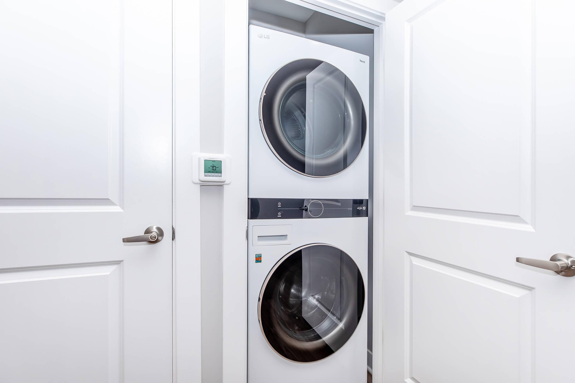 A stackable washer and dryer are installed in a small laundry area with white doors. The appliances are modern with a sleek design, and a thermostat or control panel is mounted on the wall nearby, indicating a compact and efficient use of space in a residential setting.