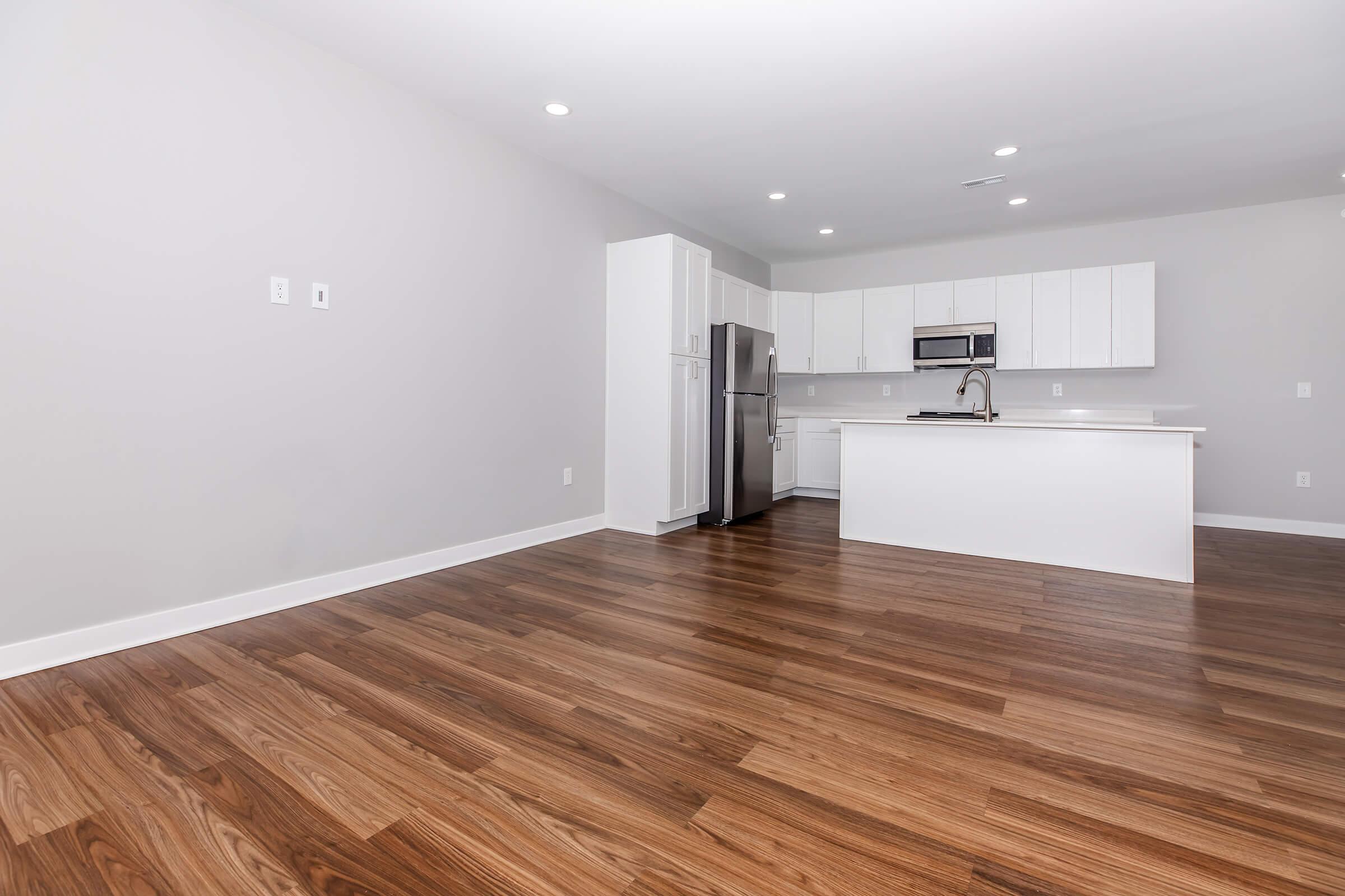 A modern kitchen with sleek white cabinetry, stainless steel appliances, and a spacious layout. The floor features rich, polished wood, and the walls are painted in a light gray color. Bright overhead lights illuminate the area, creating an open and inviting atmosphere.
