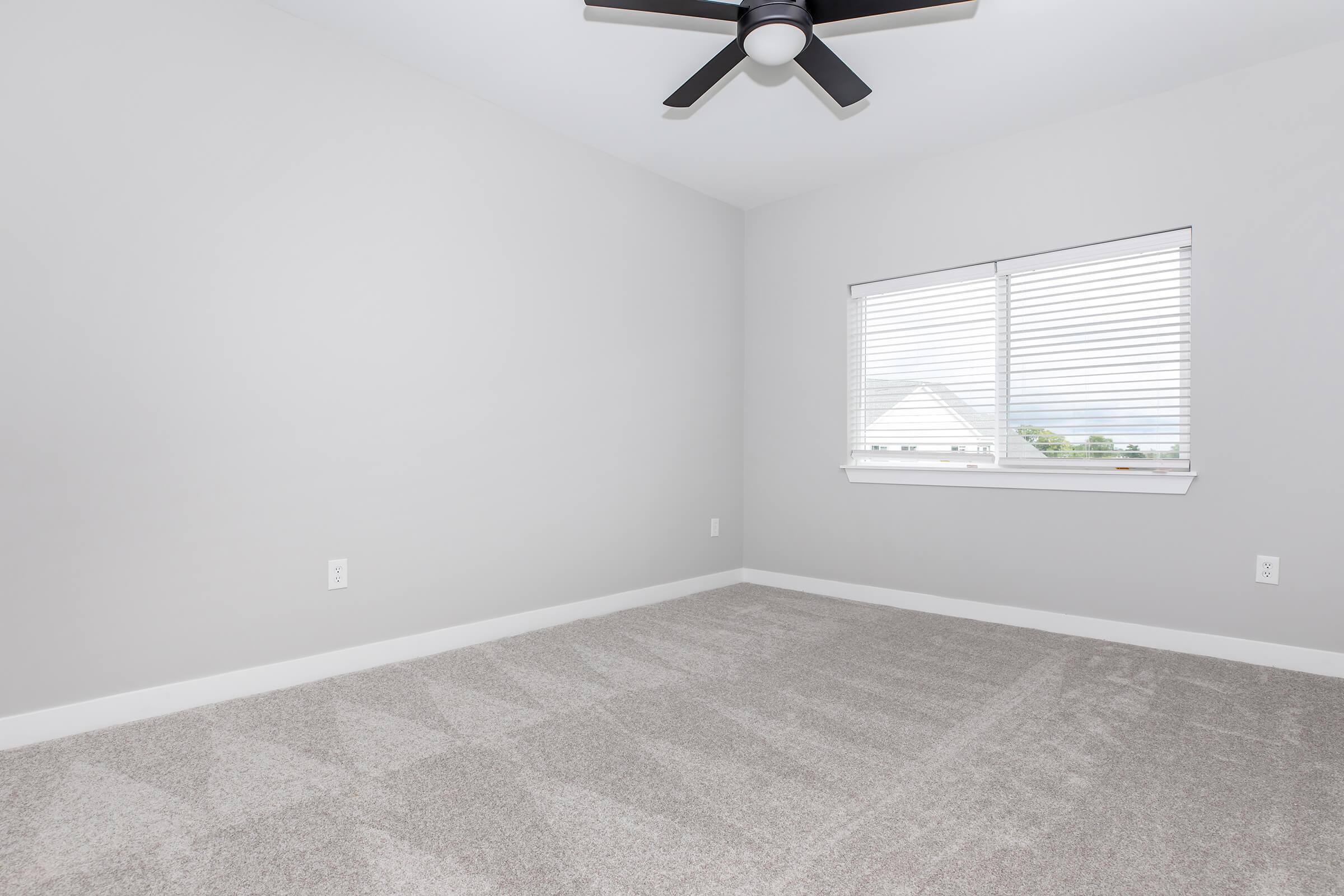 Empty room with light gray walls and carpeted flooring. A ceiling fan is mounted on the ceiling, and there is a window with white blinds letting in natural light. The room has a minimalistic and spacious feel, ideal for various uses such as an office or bedroom.