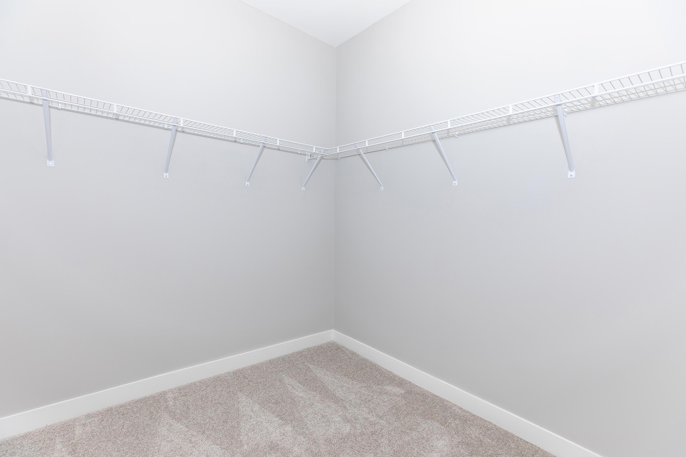 Empty closet with beige carpeting and light gray walls, featuring white wire shelving mounted on two sides. The shelving has multiple brackets but does not contain any items.
