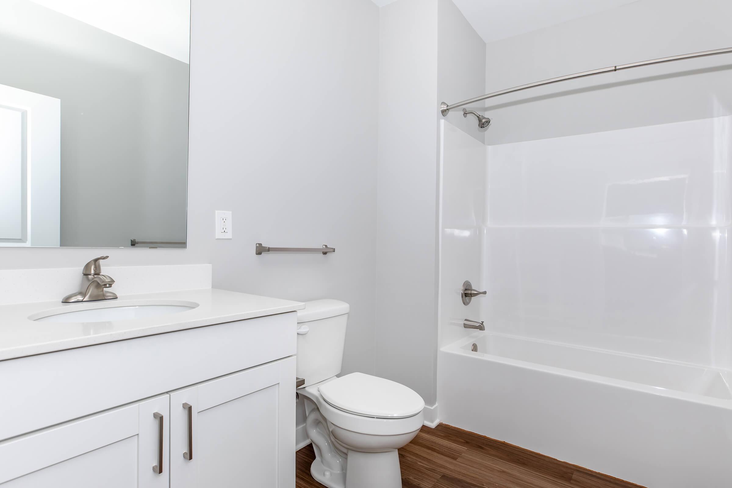 A modern bathroom featuring a white bathtub with shower, a toilet, and a vanity with a sink. The walls are painted light gray, and the floor consists of wood-like tiles. A large mirror is mounted above the vanity, and a towel bar is installed on the wall. The overall design is clean and contemporary.