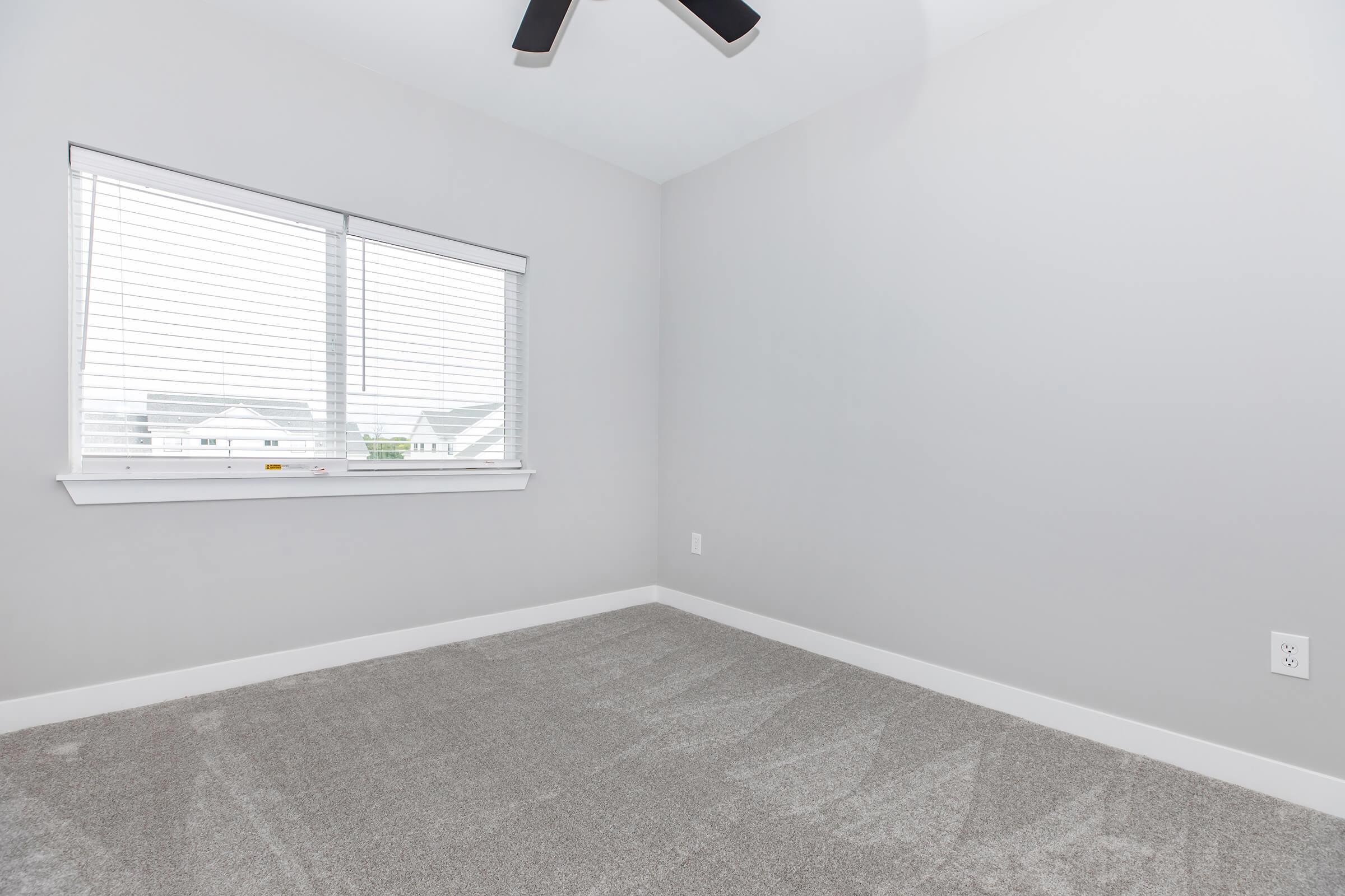 Empty room with light gray walls and a ceiling fan. There is a window with white blinds allowing natural light to enter, and the floor is covered with plush gray carpet. The space appears clean and minimalistic, suitable for various uses.