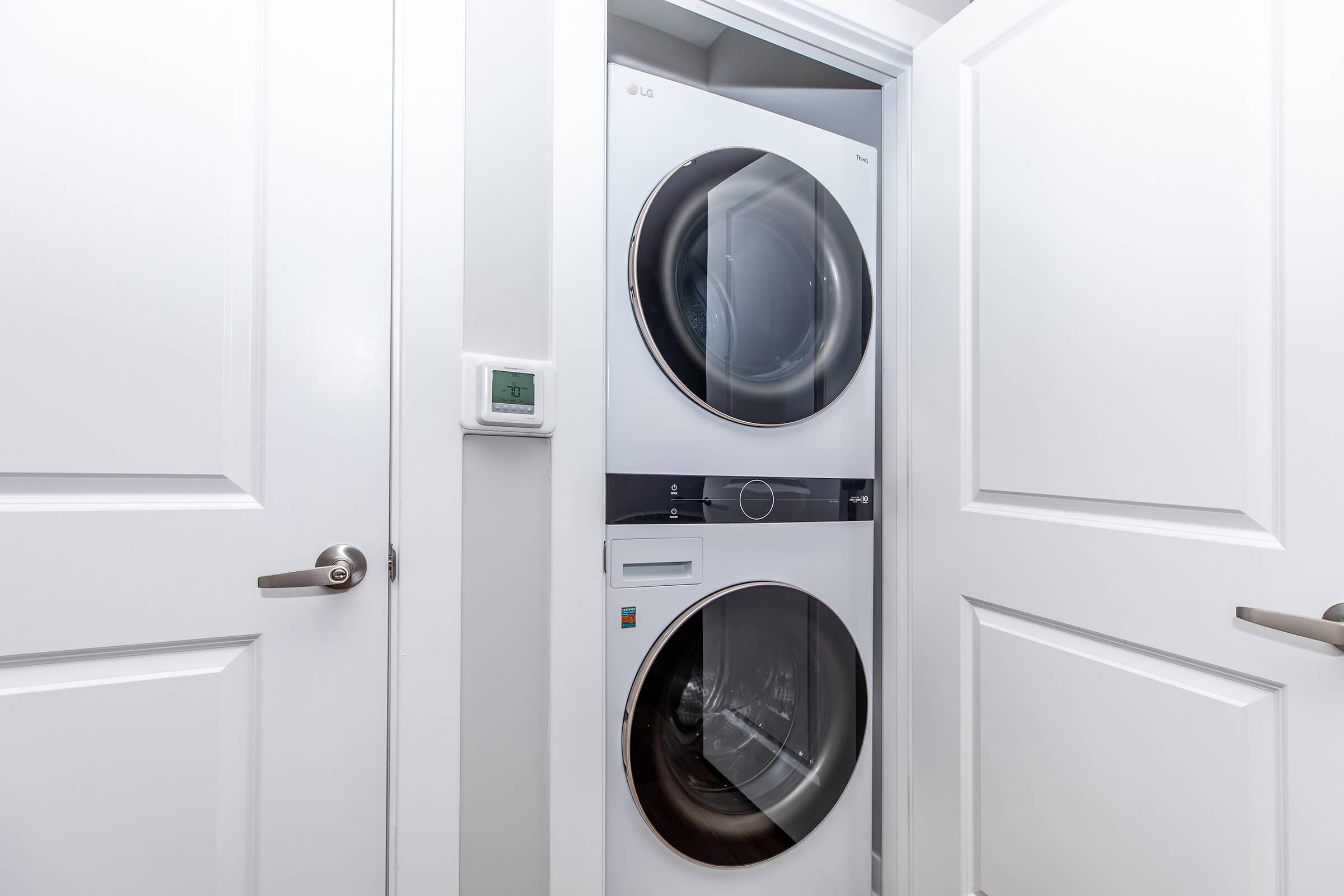 A stacked washer and dryer unit in a laundry closet, with two white doors partially open. The wall is painted white, and a small thermostat is visible next to the appliance. The overall setting appears modern and clean, suitable for integrated home laundry solutions.