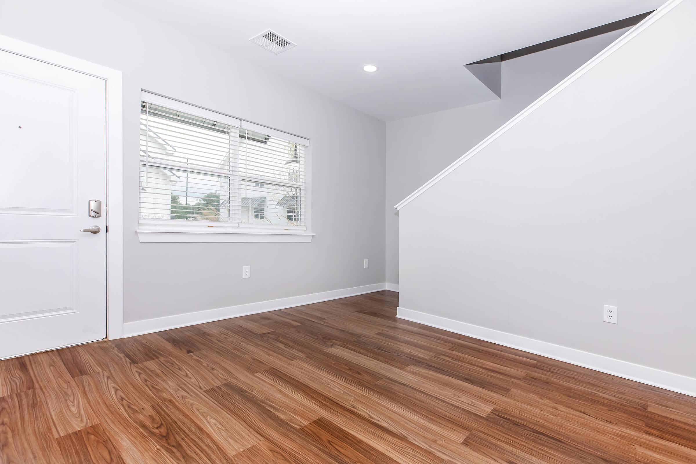 A modern interior space featuring light gray walls, a door with a small window beside it, and a staircase leading to an upper level. The floor is made of polished wood, giving a warm appearance. Natural light enters through the window, enhancing the bright and airy feel of the room.