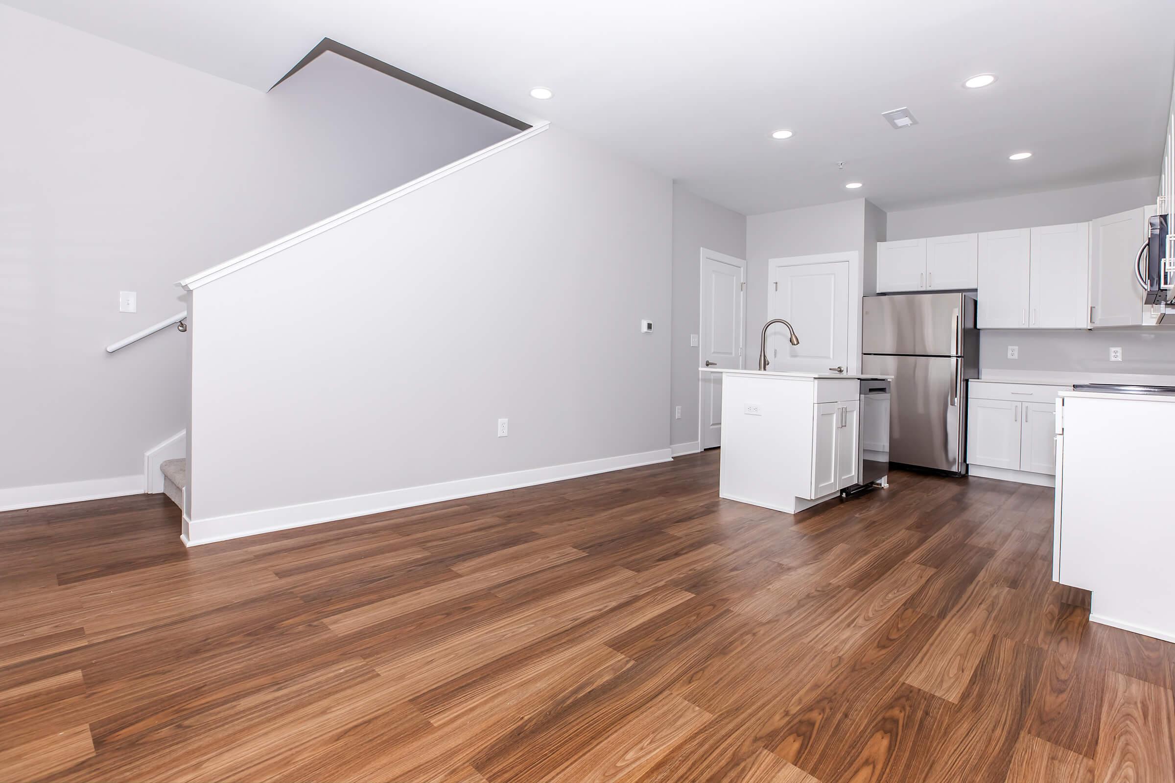 Interior of a modern kitchen and living area featuring hardwood floors, light gray walls, and a staircase. The kitchen has white cabinetry, stainless steel appliances, and a countertop island. Well-lit space with recessed lighting and a clean, open layout.