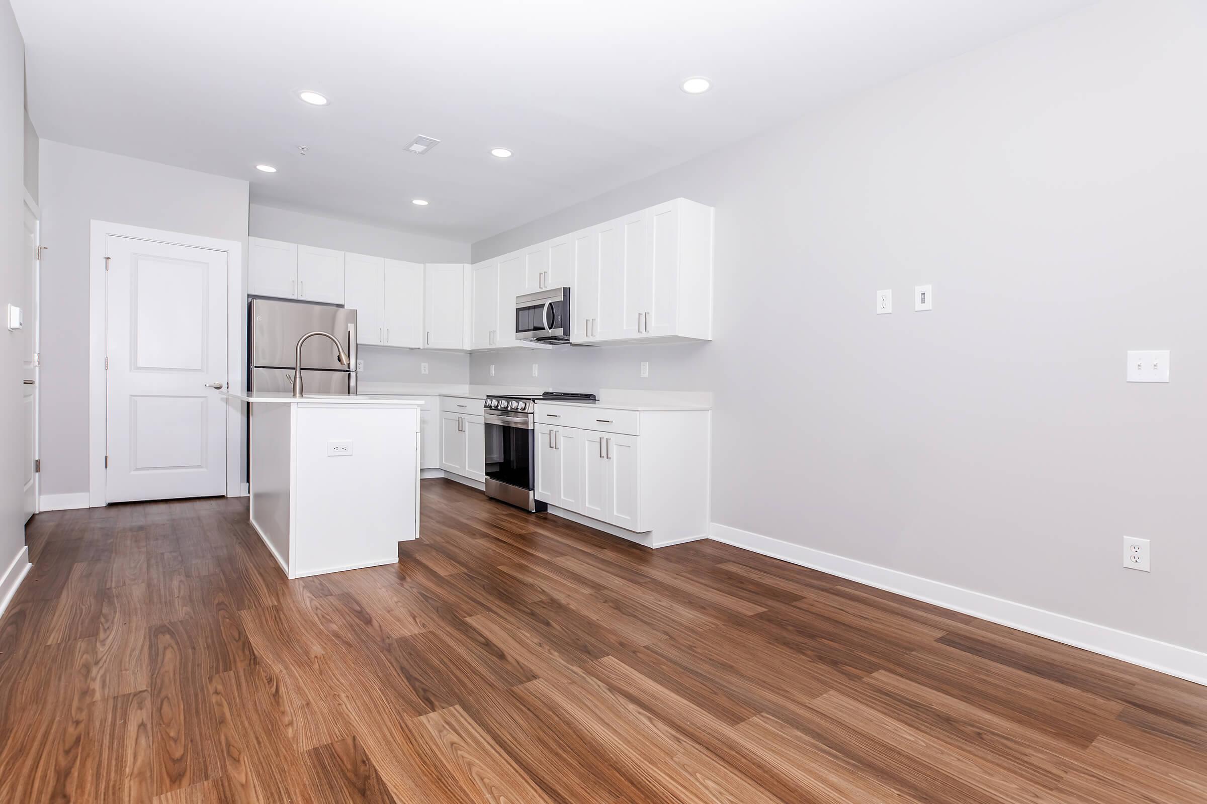 Modern kitchen featuring white cabinets, stainless steel appliances, and a large island. The flooring is a rich, dark wood, complemented by neutral gray walls and bright overhead lighting. The layout is open and spacious, enhancing the contemporary feel of the space.