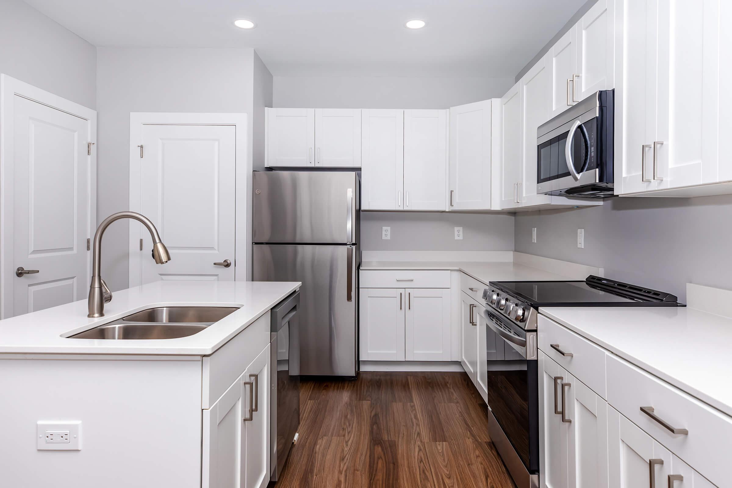 Modern kitchen featuring white cabinetry, stainless steel appliances including a refrigerator, oven, and microwave, a double sink with a sleek faucet, and wood laminate flooring. Bright and airy design with recessed lighting and a clean, minimalist layout.