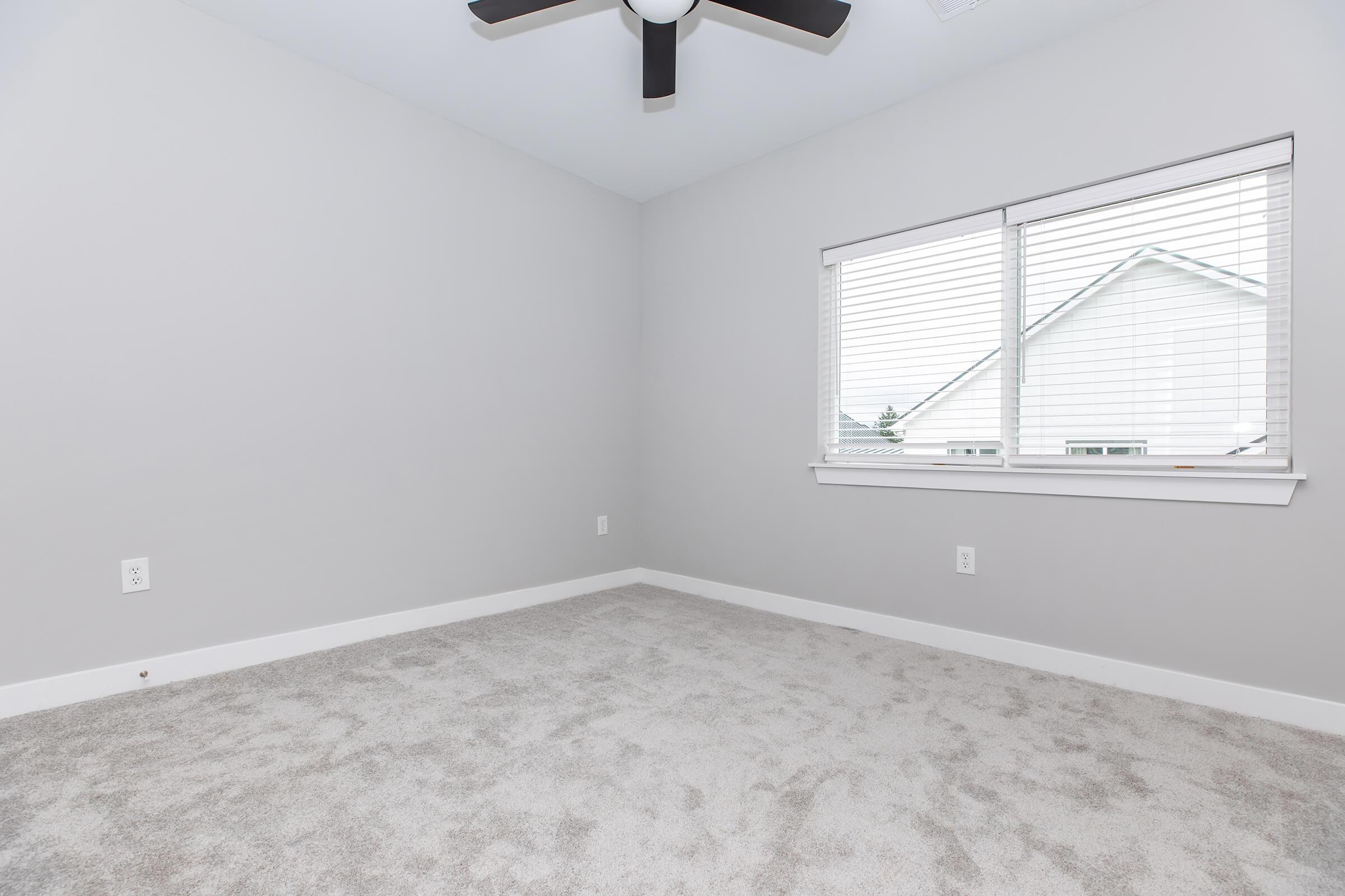 Empty room with light gray walls and carpet, featuring a ceiling fan and a window with blinds. Natural light filters through the window, which overlooks a white house outside. The space is uncluttered and ready for furniture or decoration.