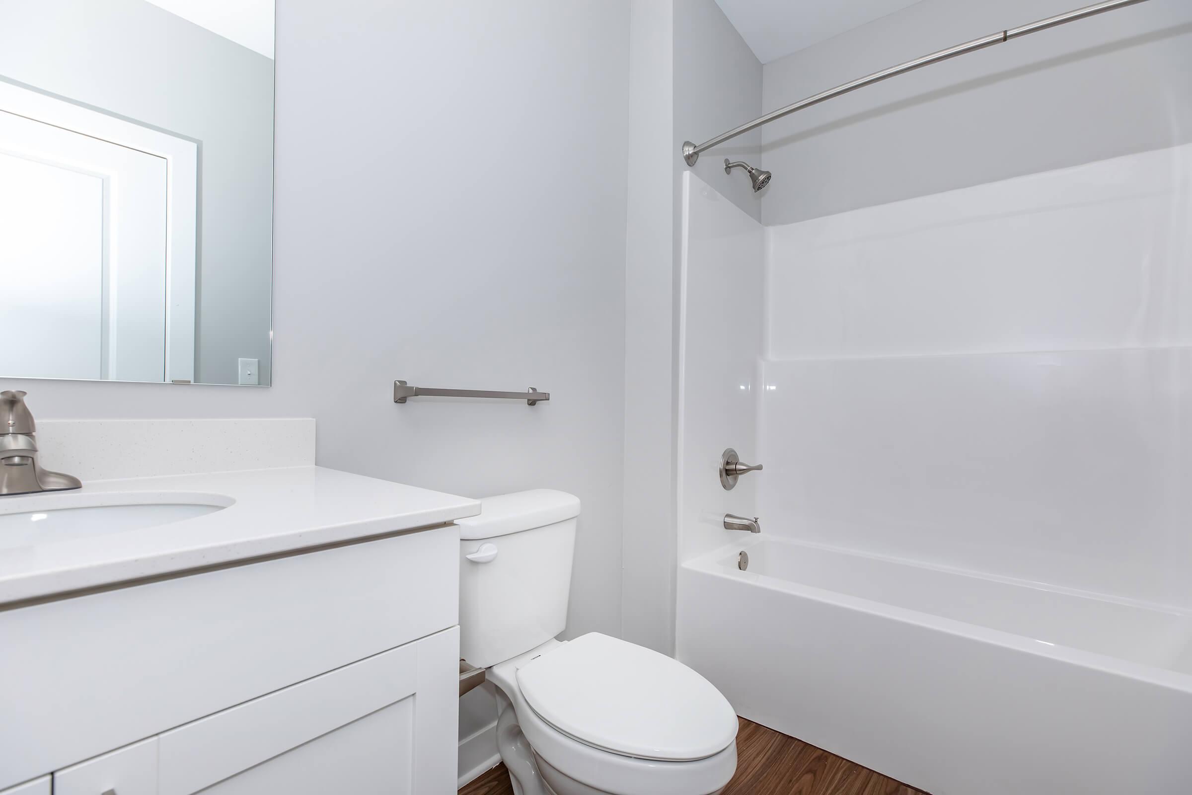 A clean and modern bathroom featuring a white bathtub with a shower rod, a toilet, and a sink with a mirror above it. The walls are painted light gray, and the floor has a wood-like appearance. The overall design is minimalistic and bright, creating an inviting space.