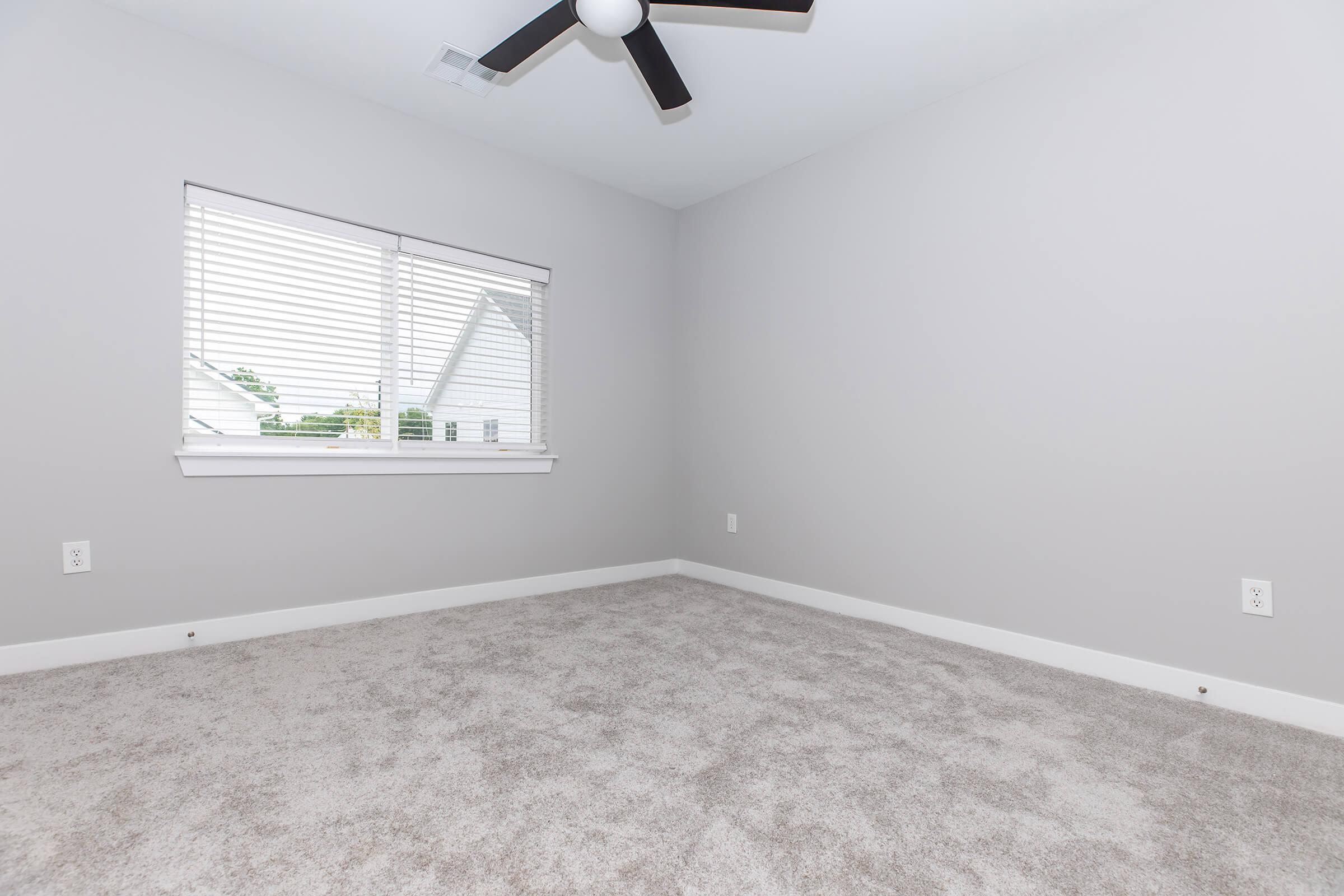 A clean, empty bedroom with light gray walls and carpet. A window with white blinds allows natural light to enter, and a ceiling fan is visible. The room has a minimalist design with no furniture, creating a spacious and airy feel.