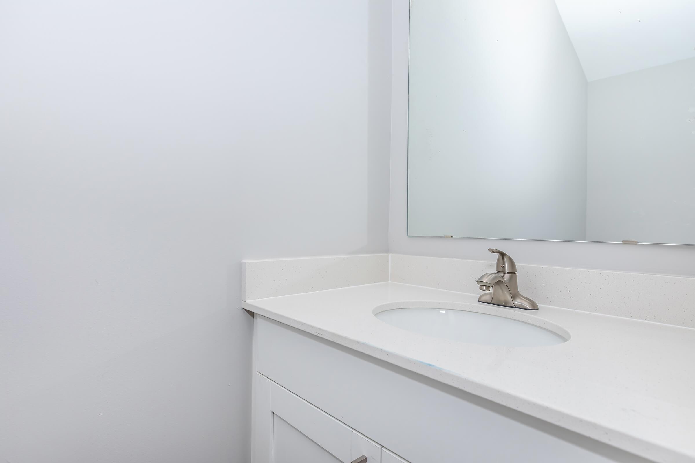 A clean, modern bathroom countertop with a stainless steel faucet and a round sink. The wall is painted white, and there is a large mirror above the sink. The overall design is minimalistic and contemporary, featuring a seamless blend of surfaces.