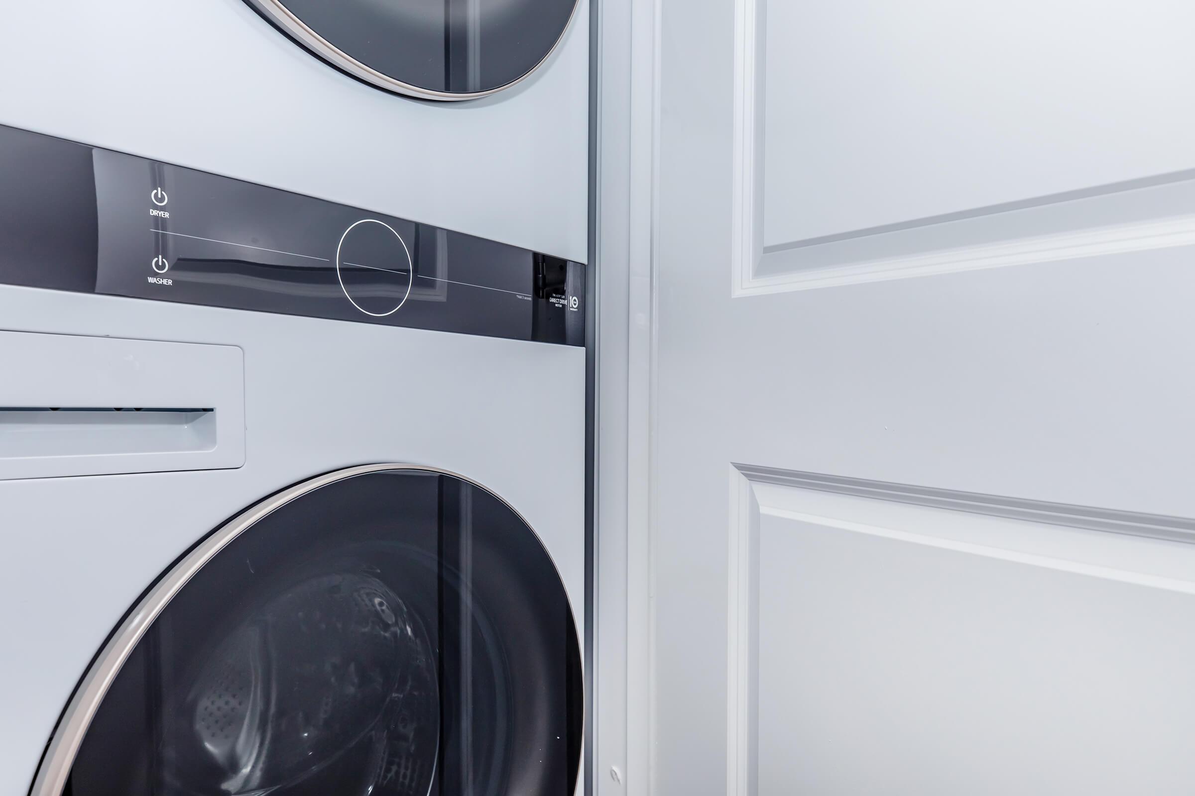 A close-up view of a stacked washer and dryer unit, with sleek, modern design features. The control panel displays various buttons and a digital display, while the door of the dryer is transparent, revealing the interior drum. The unit is positioned next to a plain white door.