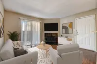 A cozy living room featuring a light-colored sofa and armchairs, a coffee table with decorative items, and a modern TV mounted on the wall. The space is complemented by a patterned rug, a large window with blinds, and artwork on the walls. A door leads outside, enhancing the inviting atmosphere.