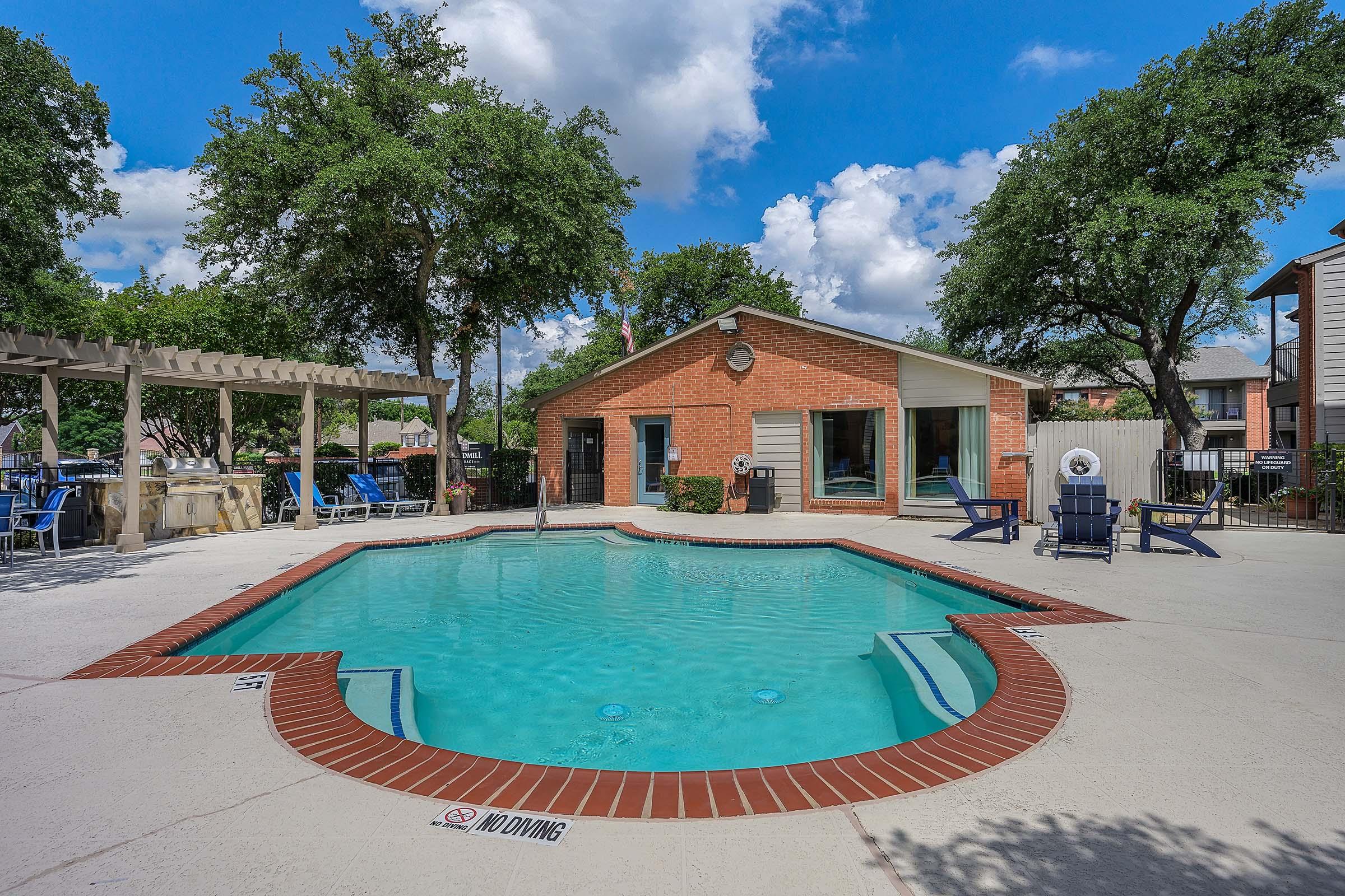 A clear swimming pool surrounded by a patio area, with lounge chairs and shaded seating. In the background, a brick house and trees under a partly cloudy blue sky. The pool has a "No Diving" sign, and the surroundings are well-maintained, creating a relaxing atmosphere.