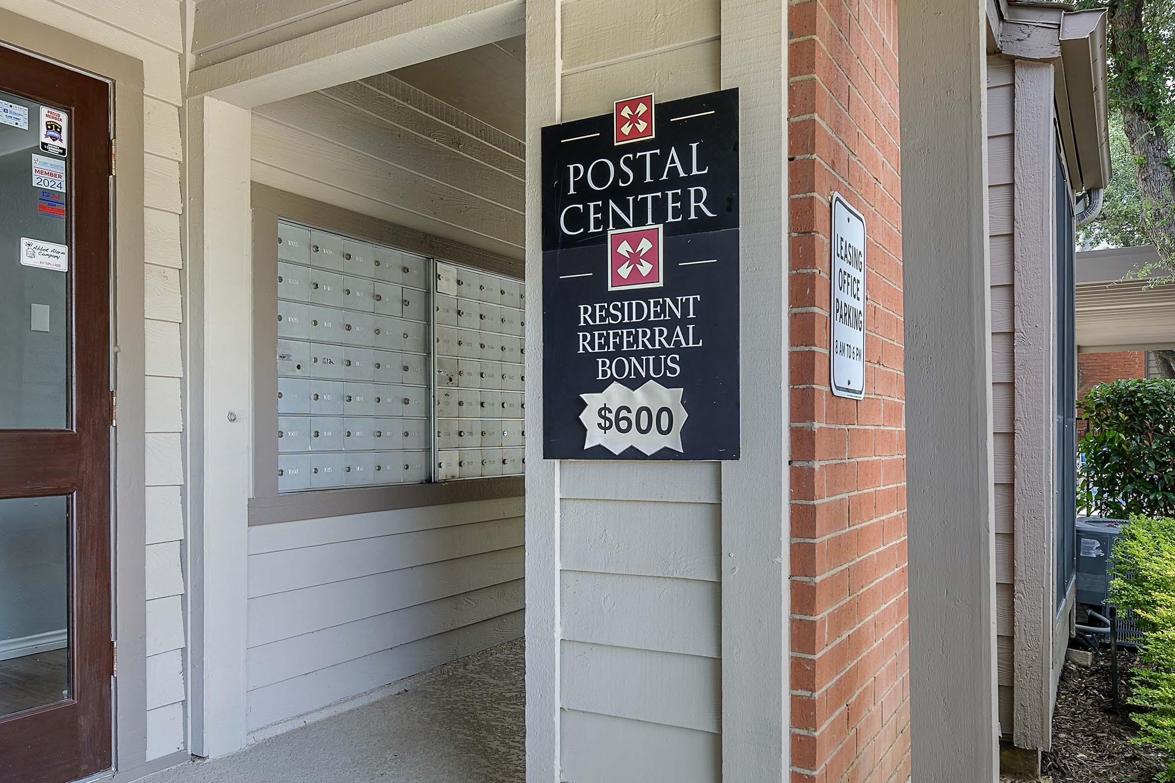 A sign for a Postal Center displaying a "Resident Referral Bonus" of $600, located next to a series of mailboxes. The entrance features a door on the left and a brick wall on the right, with greenery visible outside.
