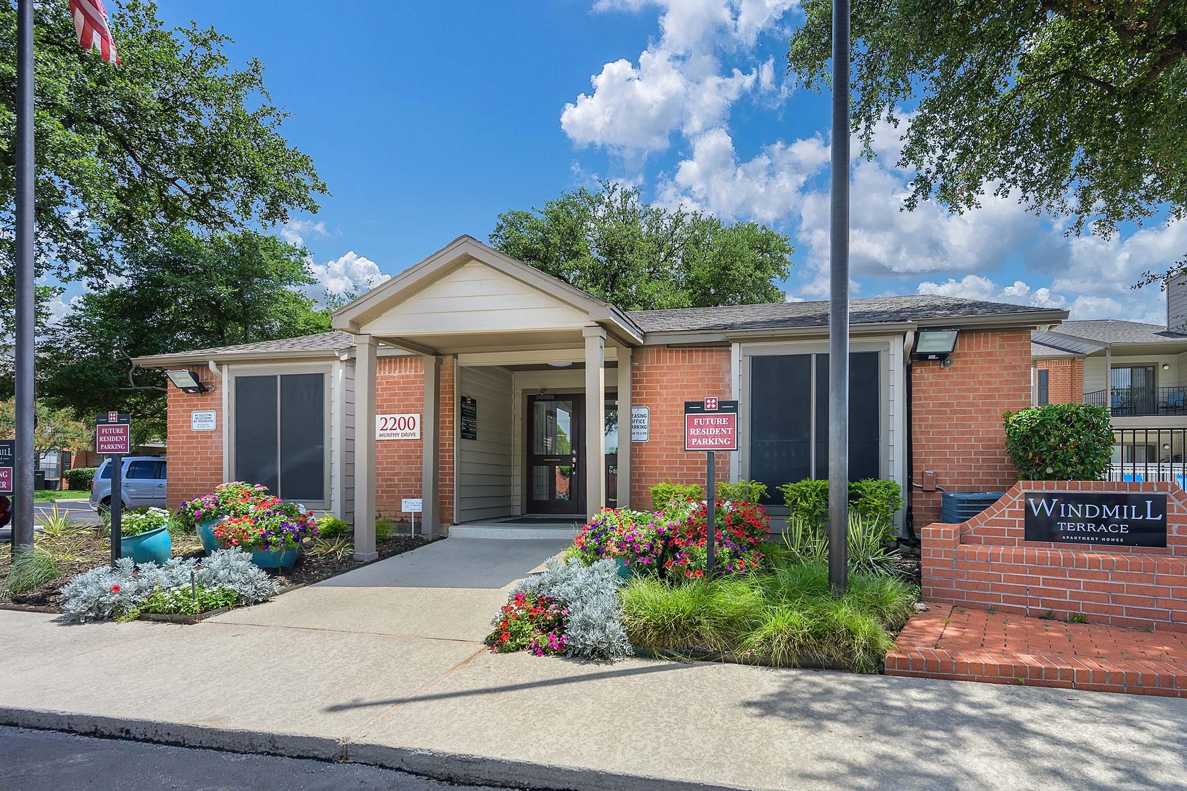 A single-story building with a beige exterior and a sloped roof, featuring large windows and a welcoming entrance. Colorful flower beds surround the entrance, and a sign reads "Windmill Terrace." The sky is mostly sunny with a few clouds.