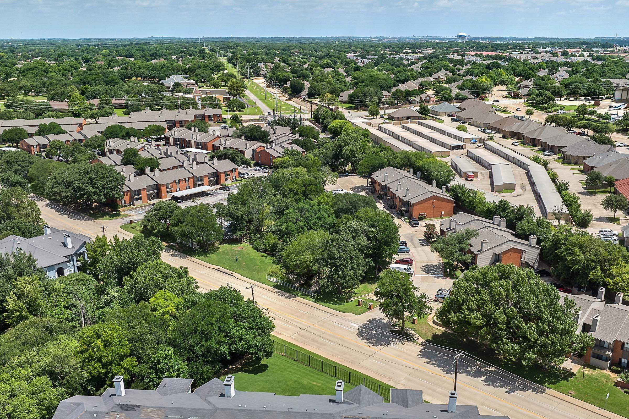 Aerial view of a suburban neighborhood featuring rows of residential buildings, lush green trees, and a winding road. Trails and greenery are visible, along with parking areas and nearby commercial buildings. The landscape is dotted with various shades of green, creating a vibrant community setting.