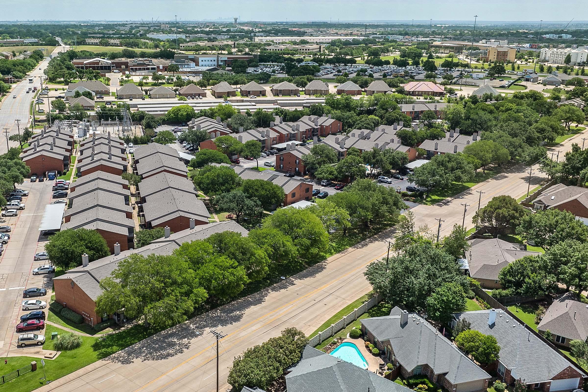 An aerial view of a suburban area featuring a mix of residential buildings, including townhouses and single-family homes, surrounded by trees. A road runs through the foreground, and several cars are parked along the streets. In the background, there are additional buildings and green spaces.