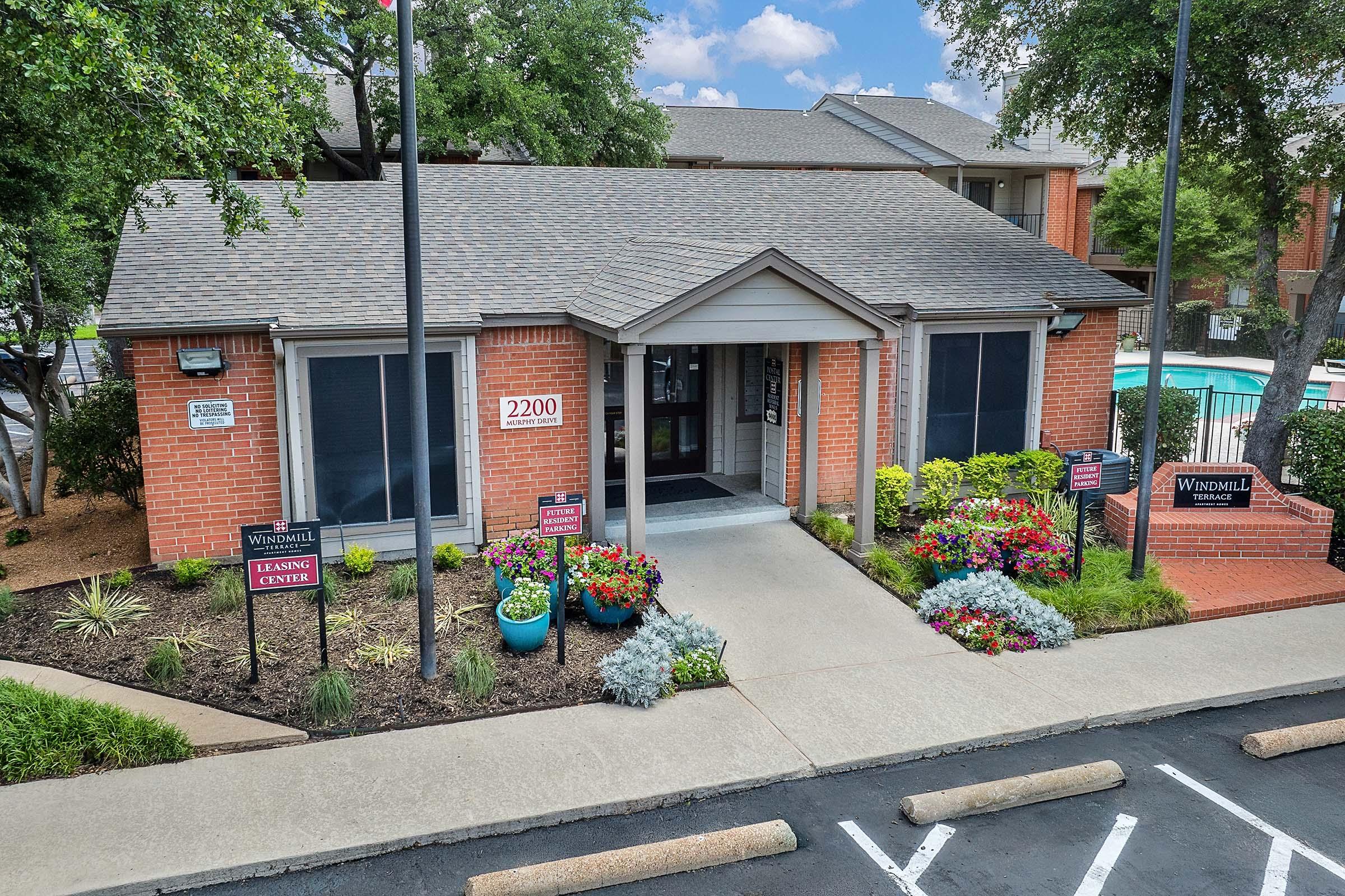 Leasing center of an apartment complex, featuring a brick facade with a gabled roof. Sign displays "2200" at the entrance. The garden is well-maintained, with colorful flower beds and potted plants. A swimming pool is visible in the background, and several parking spaces are in front.