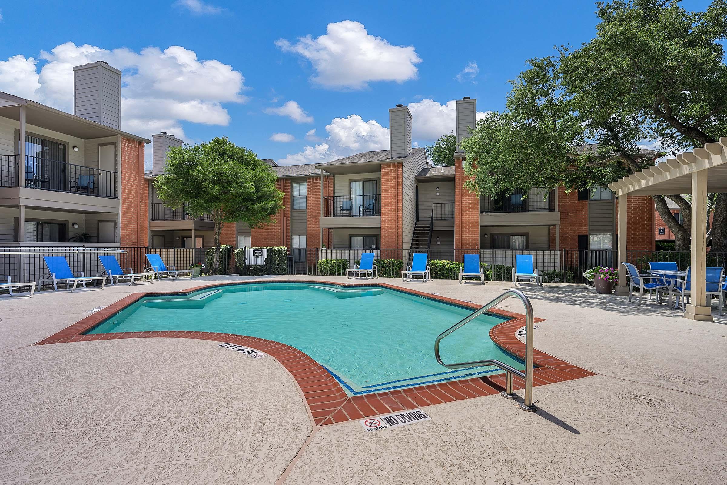 A sunny outdoor swimming pool surrounded by lounge chairs and greenery, located in the courtyard of an apartment complex. The building features multiple balconies and light-colored exteriors, with blue skies and fluffy clouds in the background.