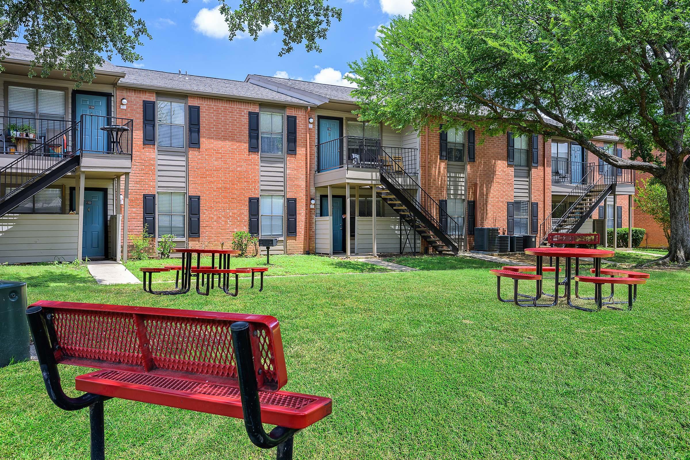 A view of an apartment complex featuring two-story buildings with red brick and light-colored siding. There are several stairs leading to the upper units and benches with tables made of metal and wood situated on a well-maintained grassy area surrounded by trees.