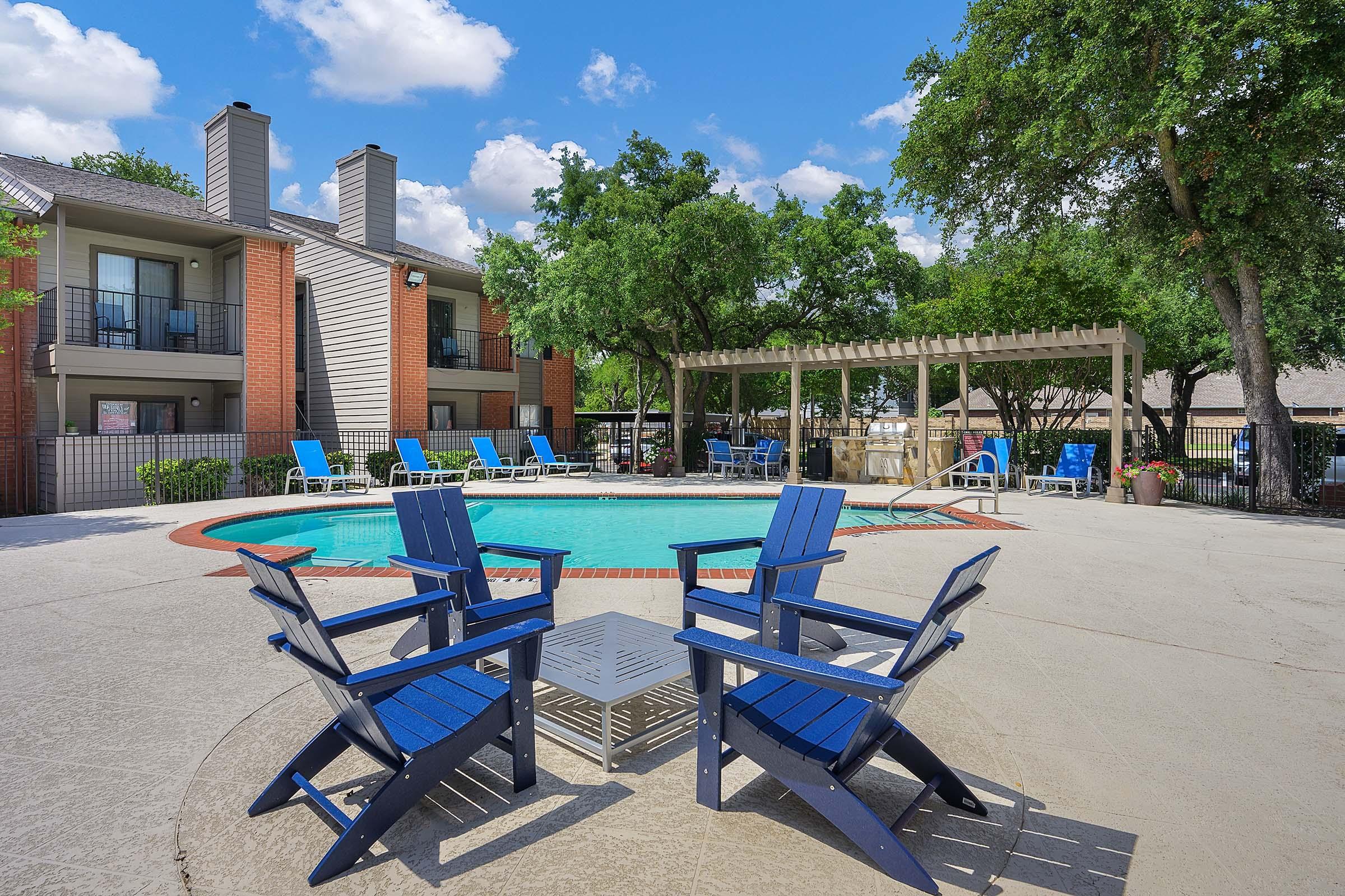 A sunny outdoor pool area featuring blue adirondack chairs around a circular pool, with white lounge chairs nearby. Tall trees provide shade, and there is a pergola in the background. The setting is inviting and perfect for relaxation.