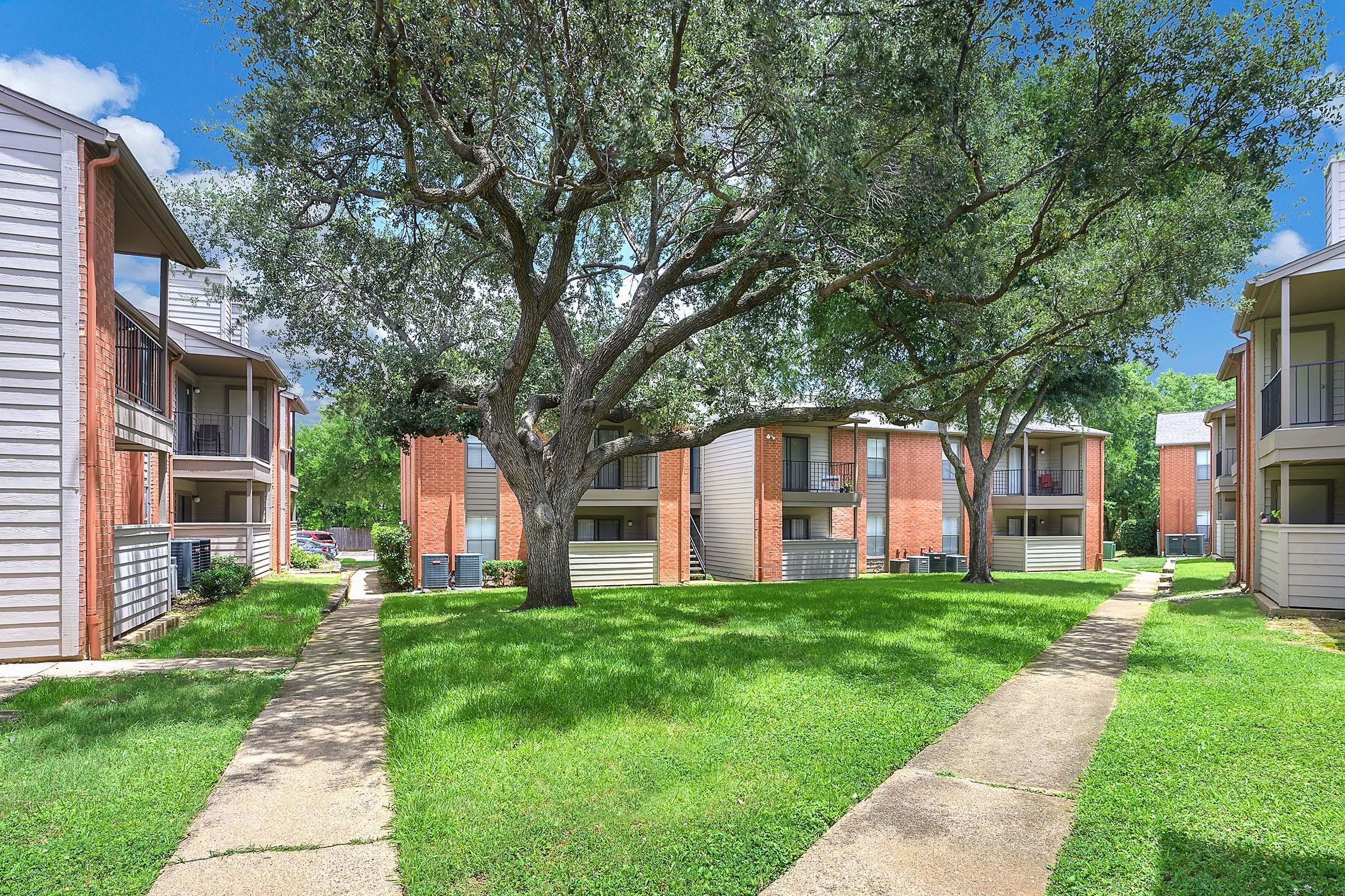 View of a residential complex featuring several two-story buildings arranged around a grassy courtyard. The area is shaded by large trees, with pathways leading through the greenery. Clear blue skies and lush lawns create a welcoming atmosphere.