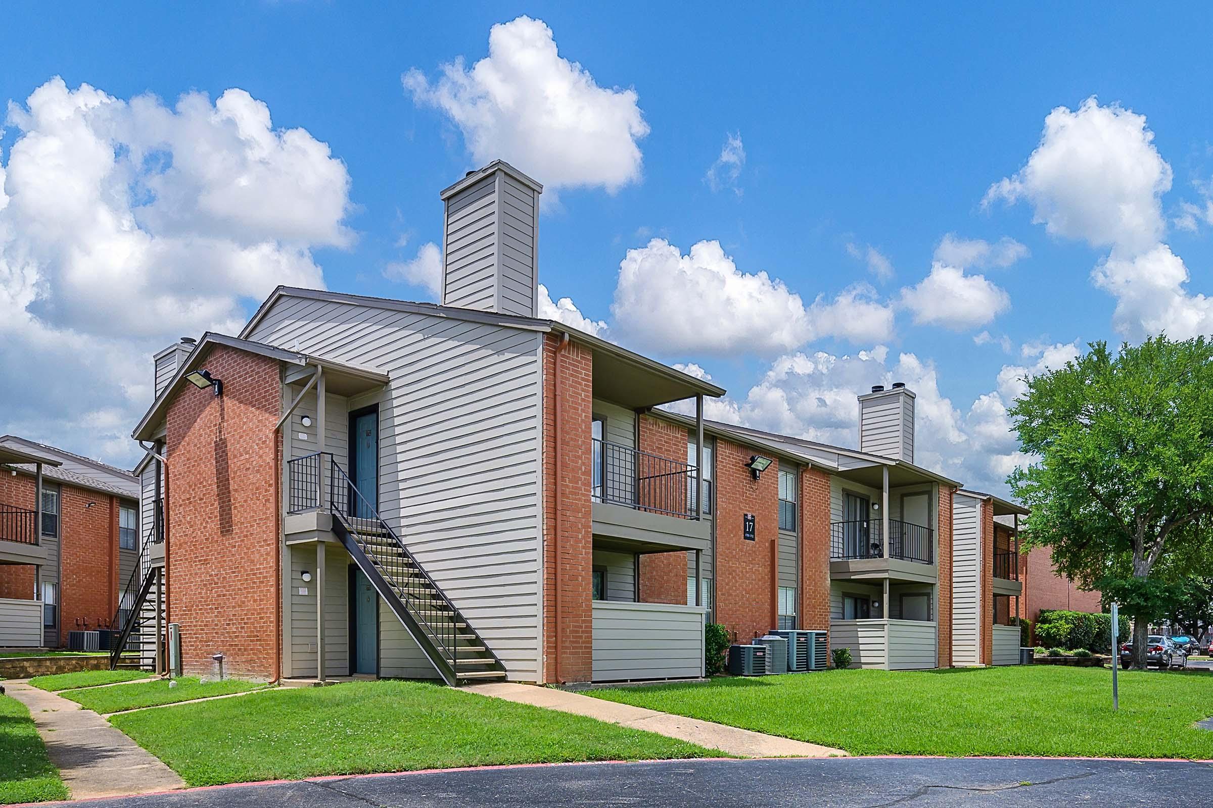 Two multi-unit apartment buildings with two stories, featuring a mix of brick and siding exteriors. Each building has exterior staircases leading to balconies. The scene includes a well-maintained green lawn, paved walkways, and a partly cloudy blue sky in the background.