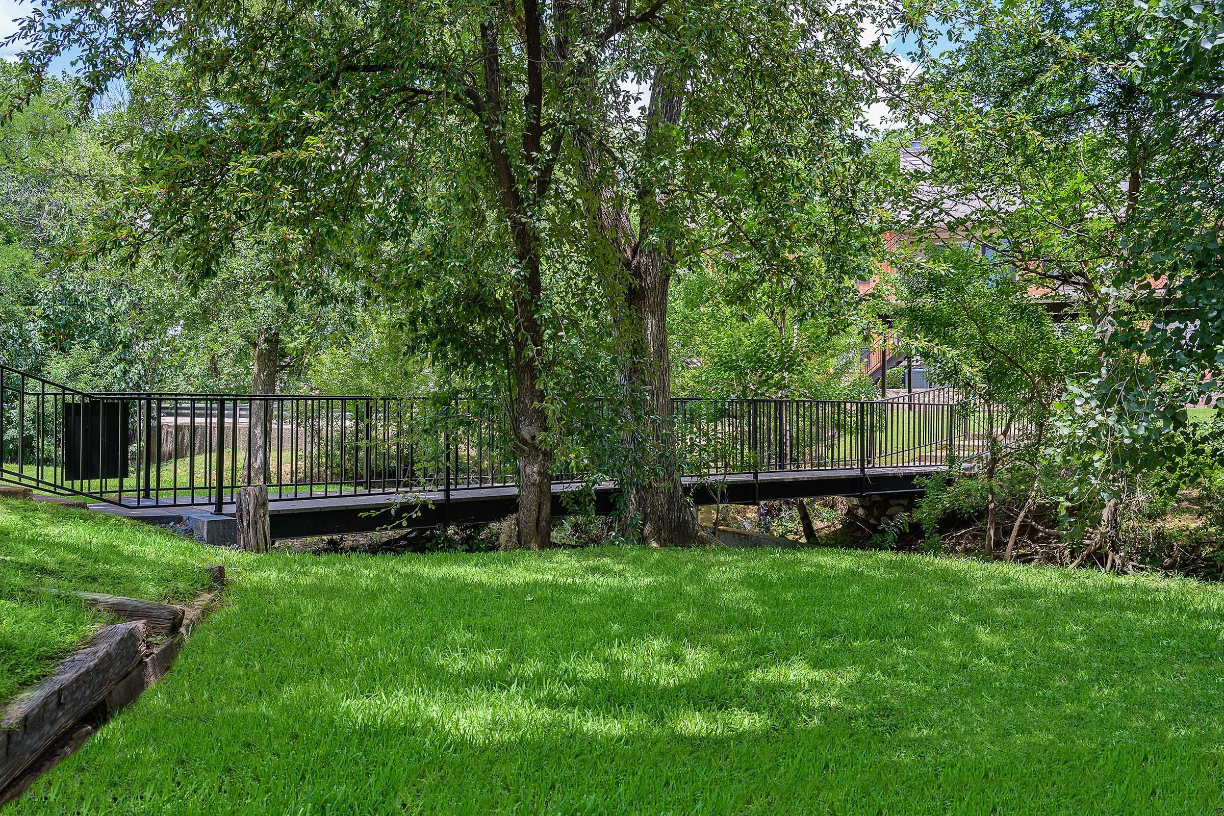 A peaceful scene featuring a metal bridge spanning a small creek, surrounded by lush greenery and trees. The well-maintained lawn in the foreground enhances the natural beauty of the area, with sunlight filtering through the leaves on a clear day.
