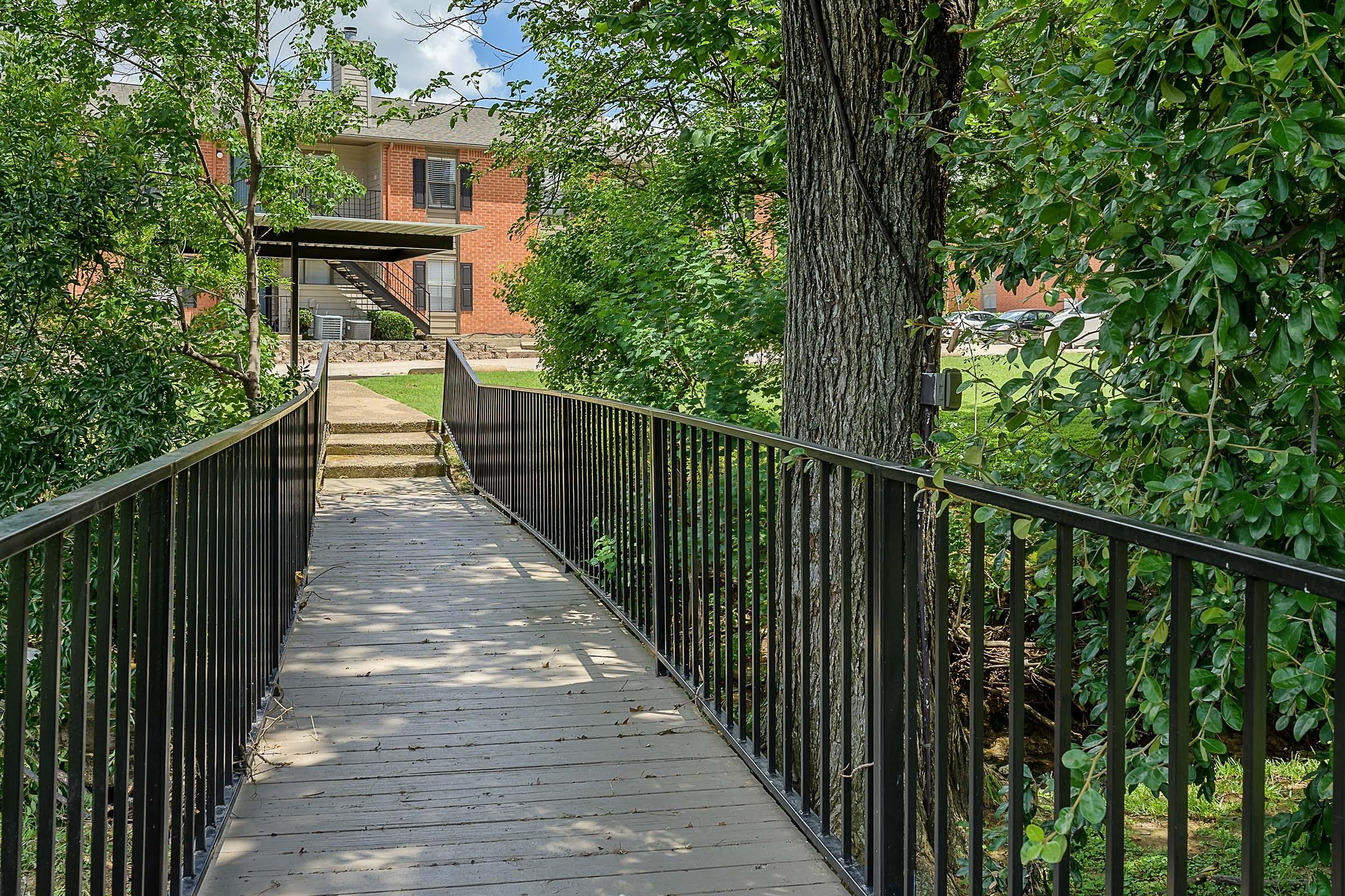 A wooden bridge with a black railing leading to a pathway surrounded by greenery. In the background, there is a red brick building partially visible, along with a shaded area beneath an awning. The scene is bright and inviting, highlighting a peaceful outdoor environment.