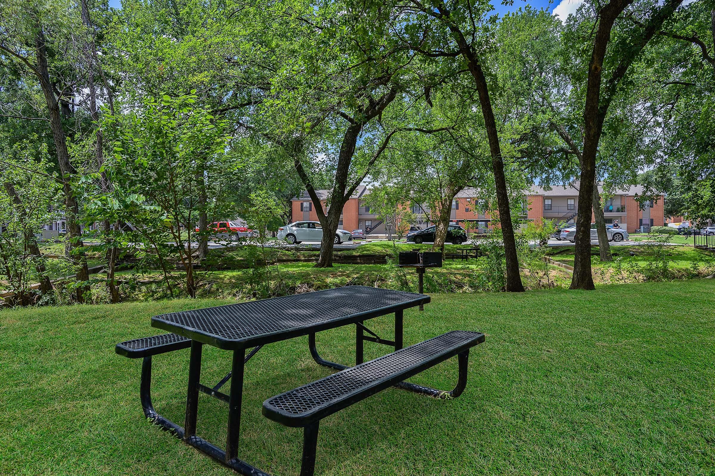 A black metal picnic table with attached benches sits on a well-maintained grassy area surrounded by trees. In the background, a row of buildings can be seen, along with parked cars and a clear blue sky overhead. The scene suggests a peaceful outdoor space for relaxation and gatherings.
