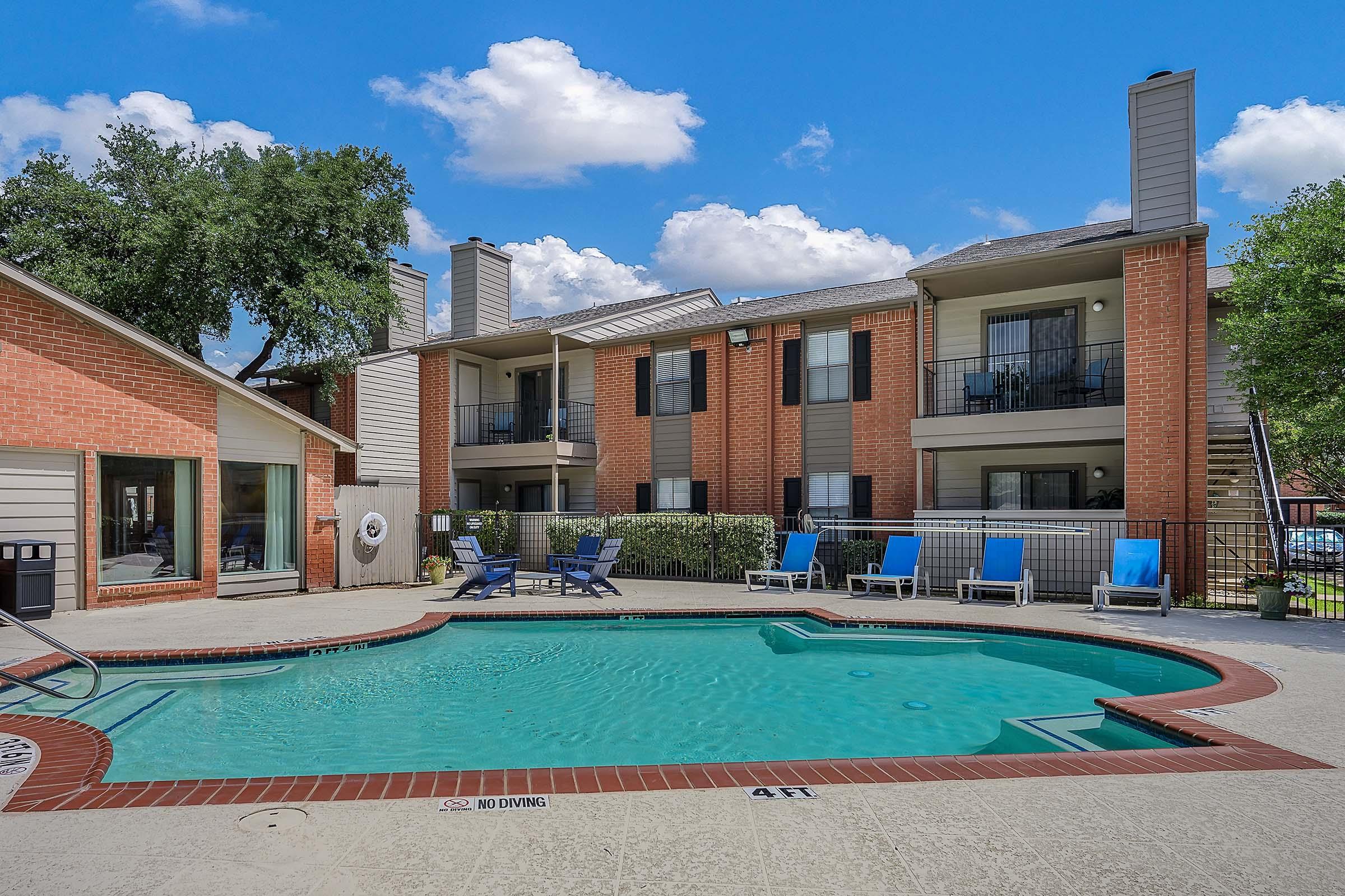 A serene apartment complex featuring a sparkling blue swimming pool surrounded by lounge chairs. The two-story buildings have a warm brick exterior, and lush greenery enhances the inviting atmosphere under a clear blue sky with scattered clouds.