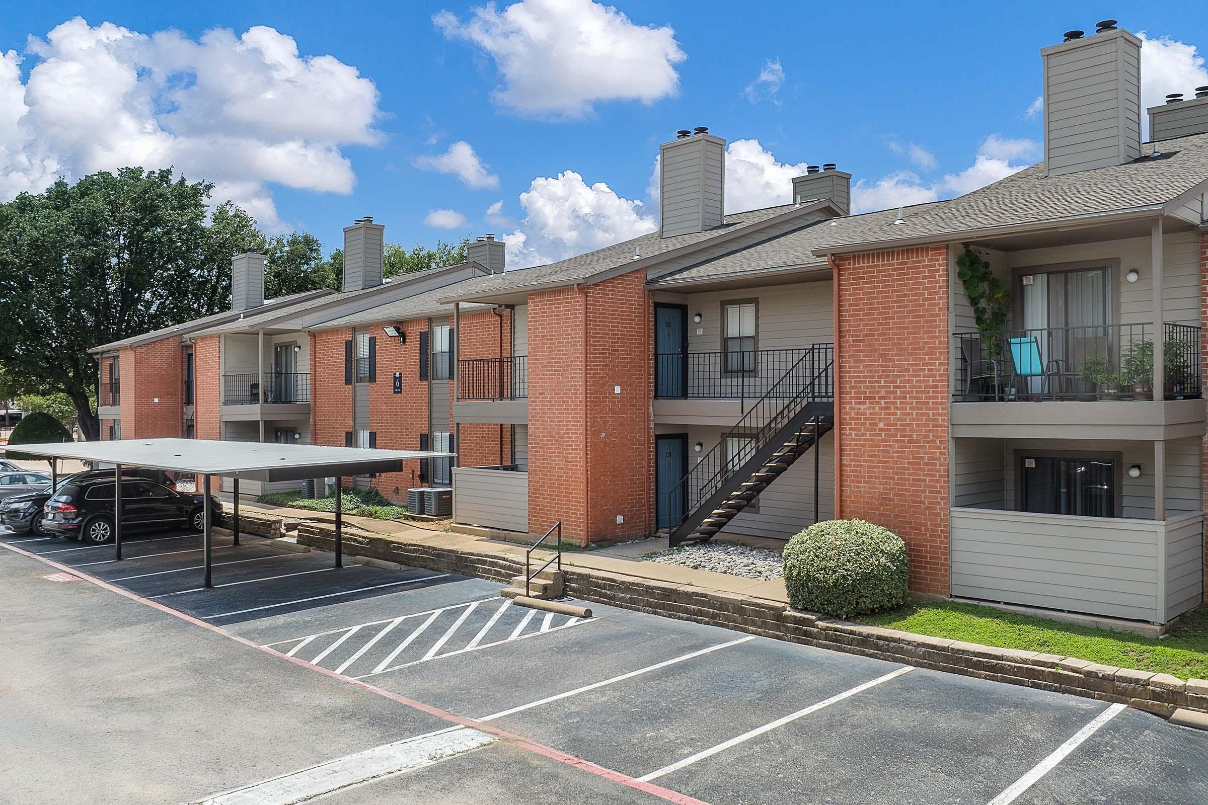 View of a multi-unit apartment building with a brick and light-colored exterior. There are parked cars in a covered parking area, and stairs leading to upper-level apartments. The setting has a clear blue sky with fluffy clouds and greenery in the background.